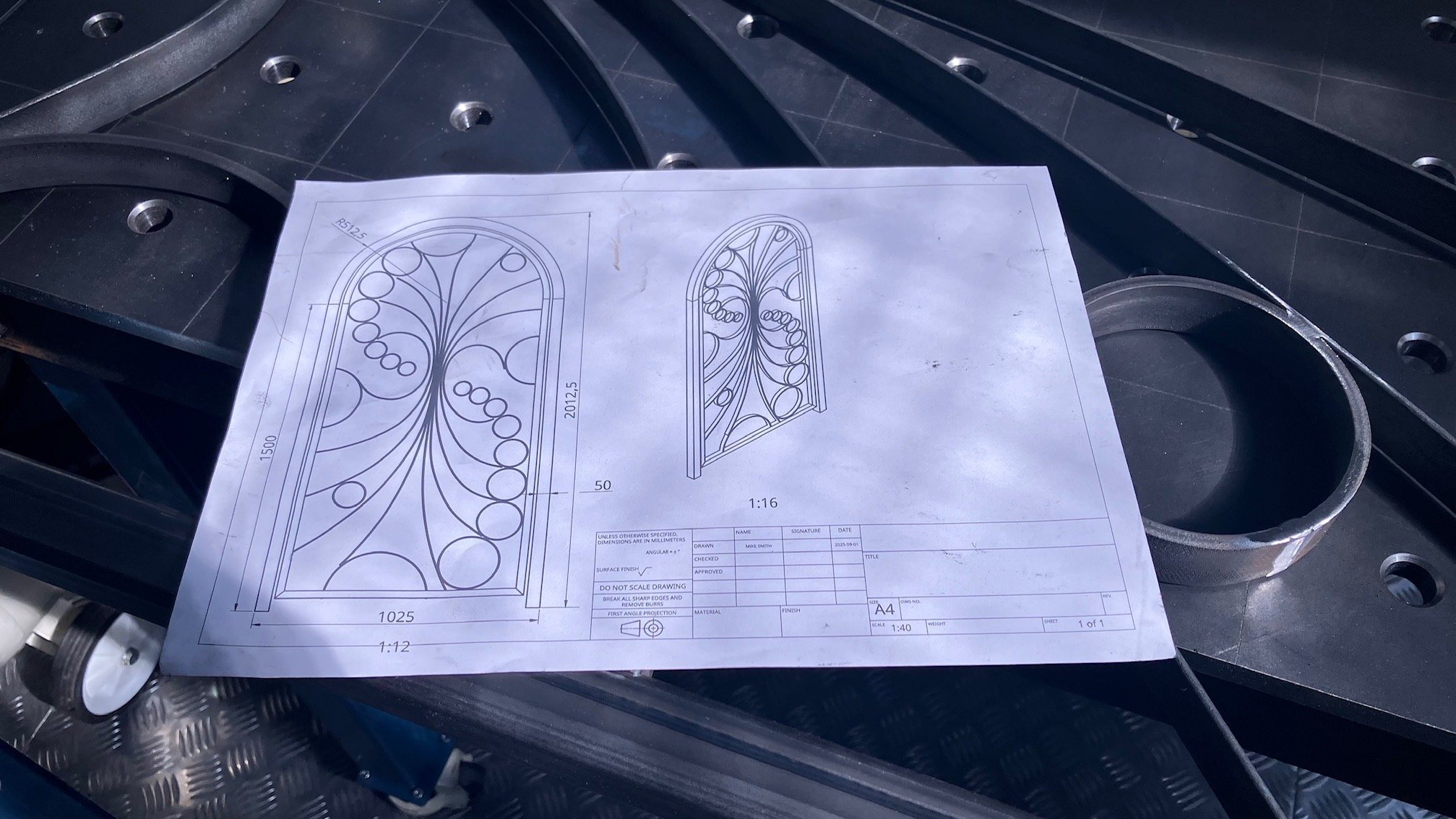 Technical drawing of a decorative window design placed on a metal work surface among metal parts.