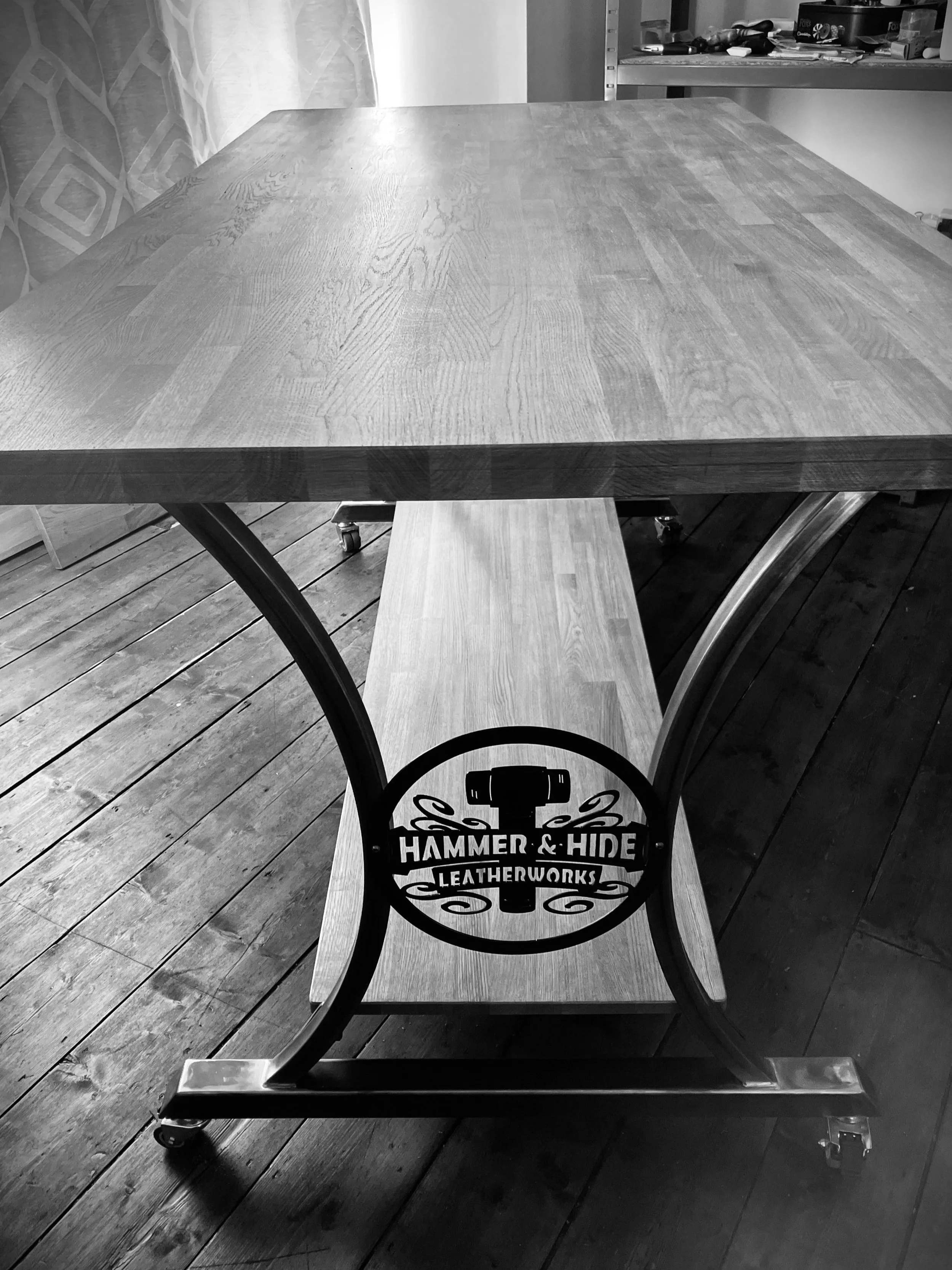 Black and white photo of a wooden table on casters, with a decorative metal sign that reads "Hammer & Hide Leatherworks," and a shelf with various tools and materials in the background.