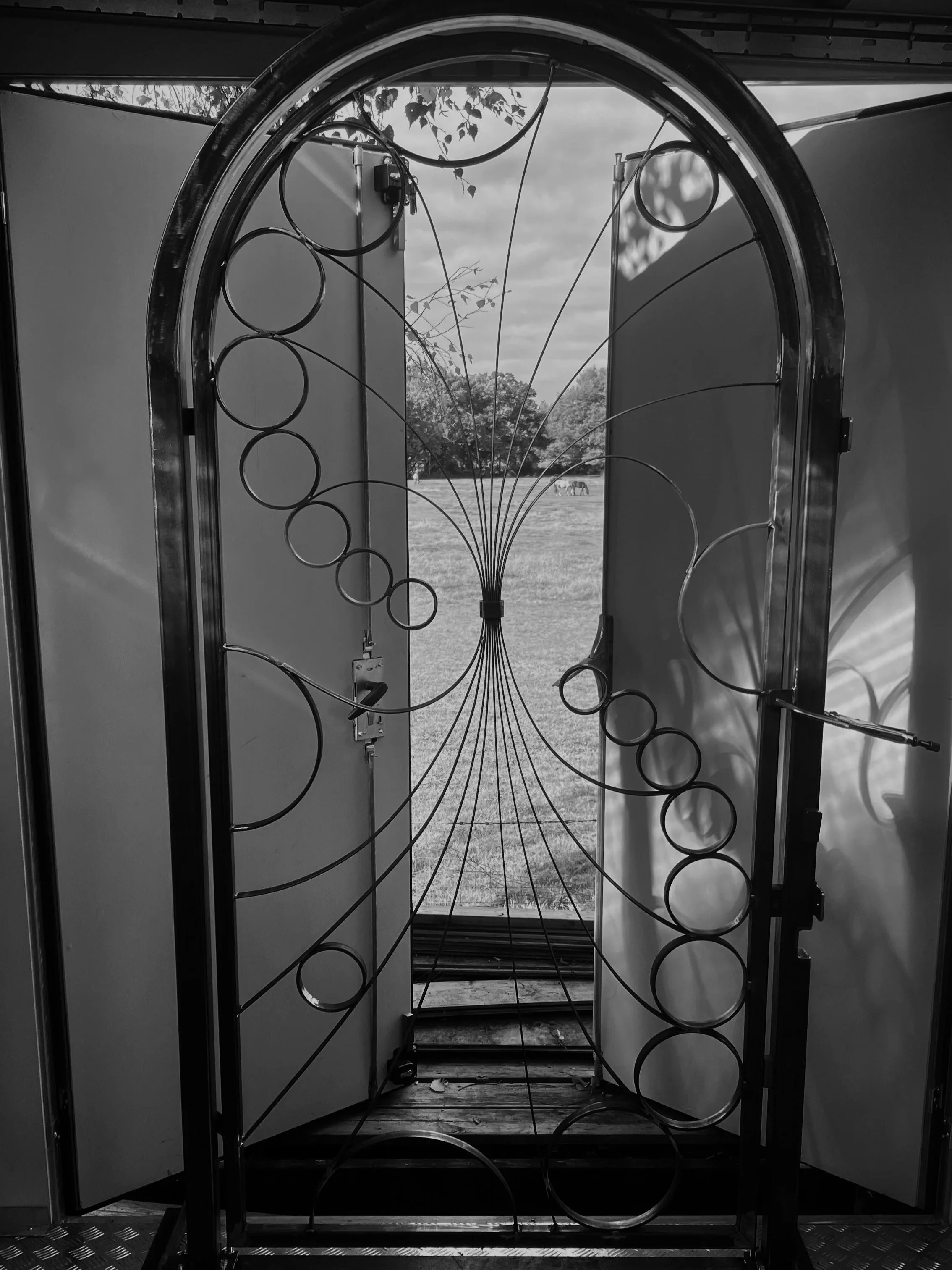 Black and white photo of a sculpture or installation visible through a decorative metal gate with circular and curved patterns, with a field, trees, and horses in the background.