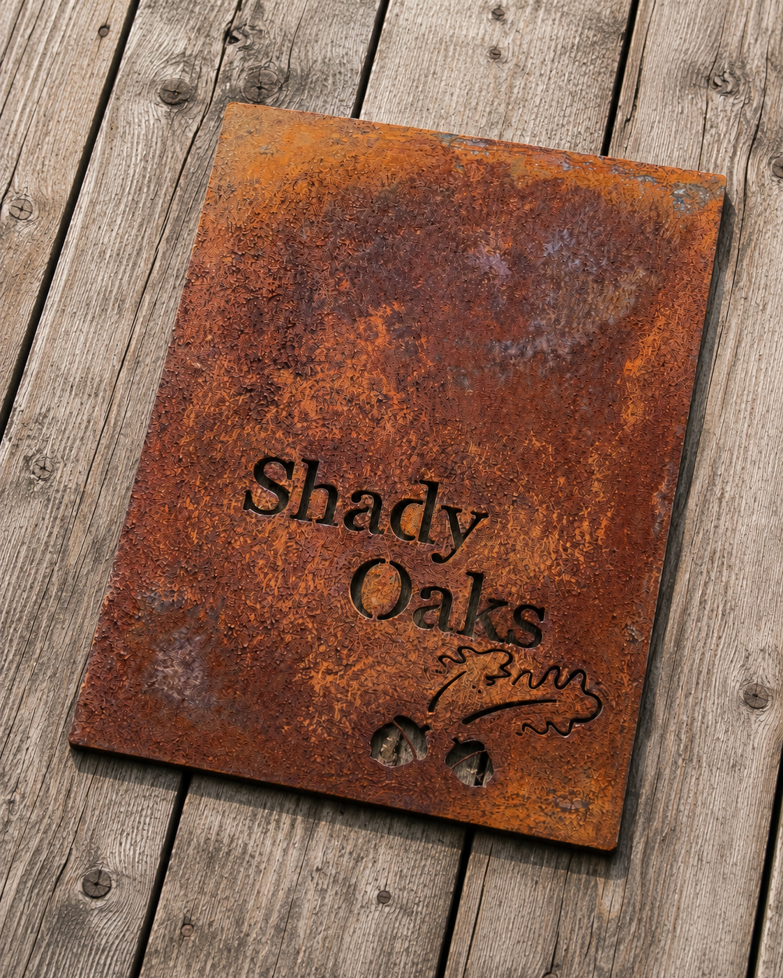 Rusty sign with cut-out text reading 'Shady Oaks' and a tree branch illustration, placed on weathered wooden planks.