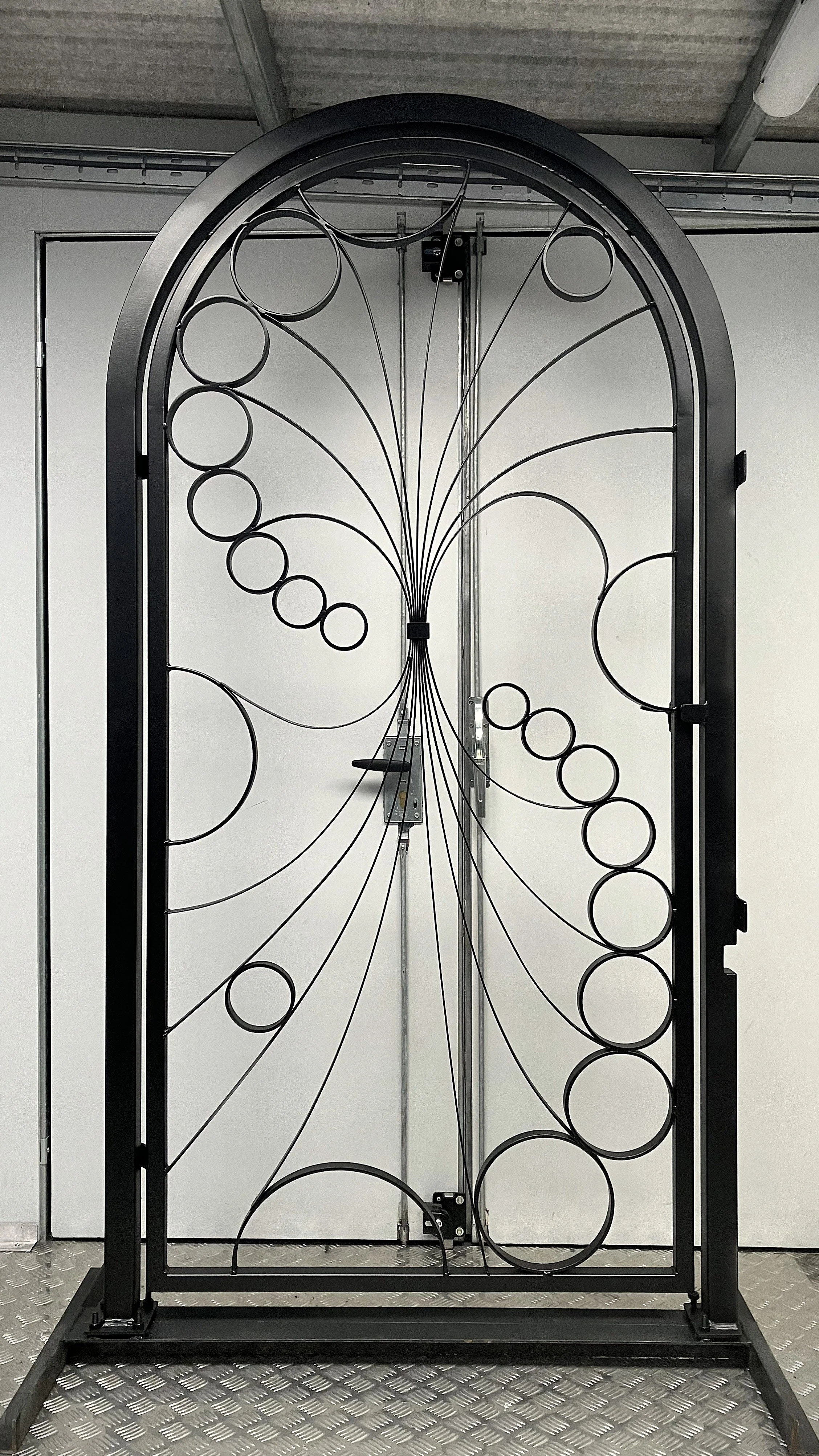 A decorative metal gate with circular patterns, black frame, standing in an industrial or warehouse space with gray walls and ceiling.
