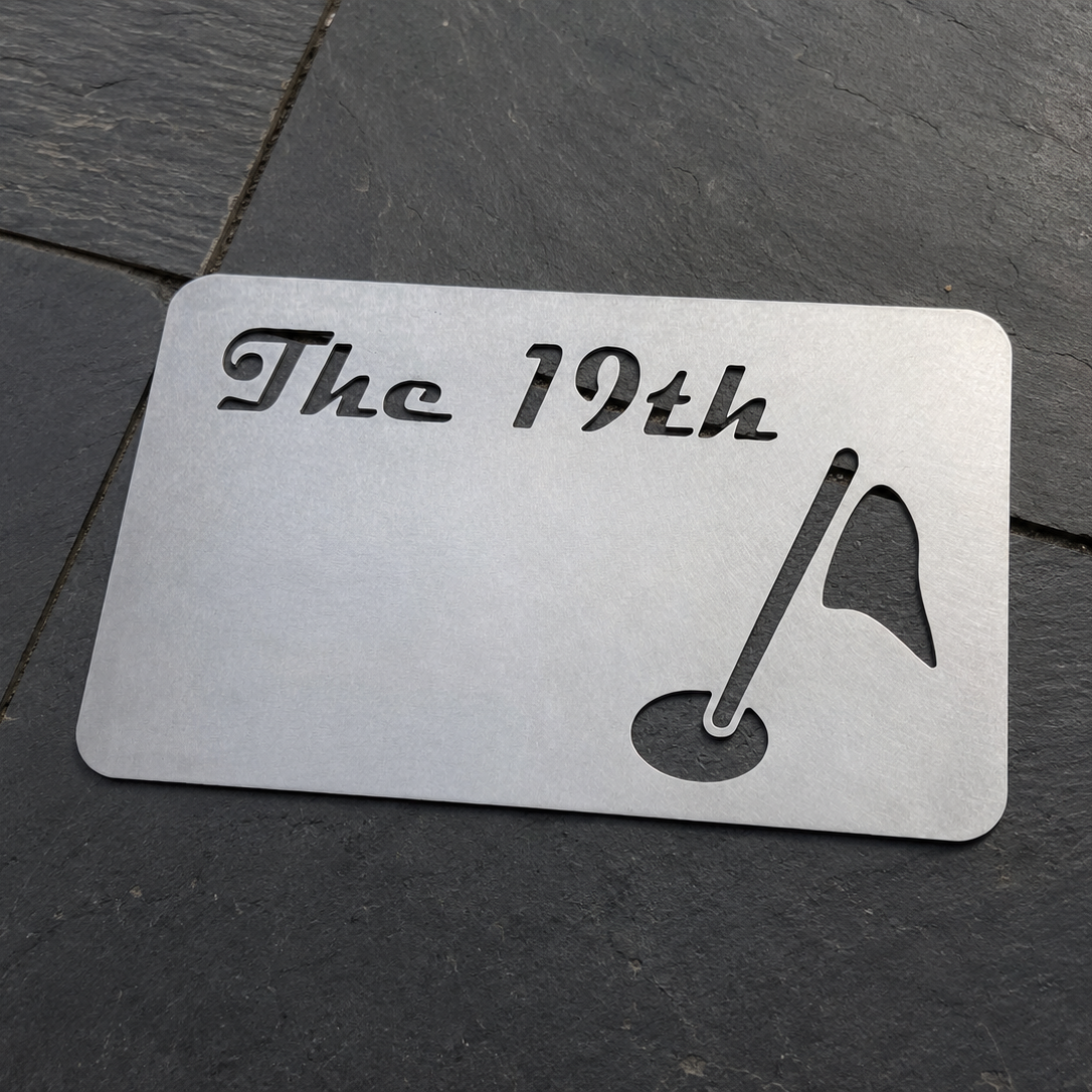 A metallic cut-out sign with the text 'The 19th' and a golf flag and hole illustration, placed on a gray stone surface.