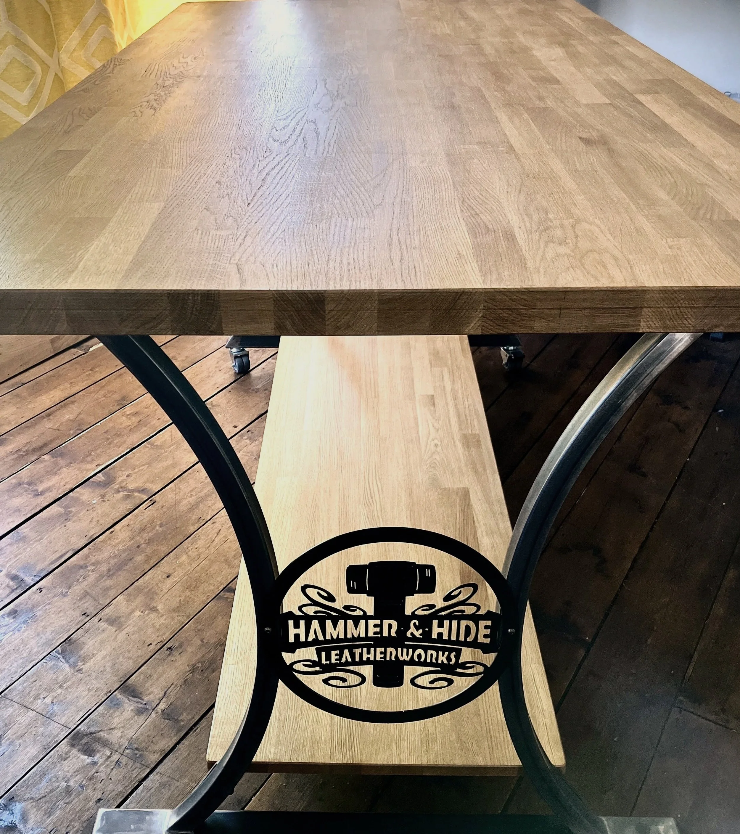 A steel legged table with a black metal sign that reads 'Hammer & Hide Leatherworks', placed in a space with wooden plank flooring and a yellow patterned wall.