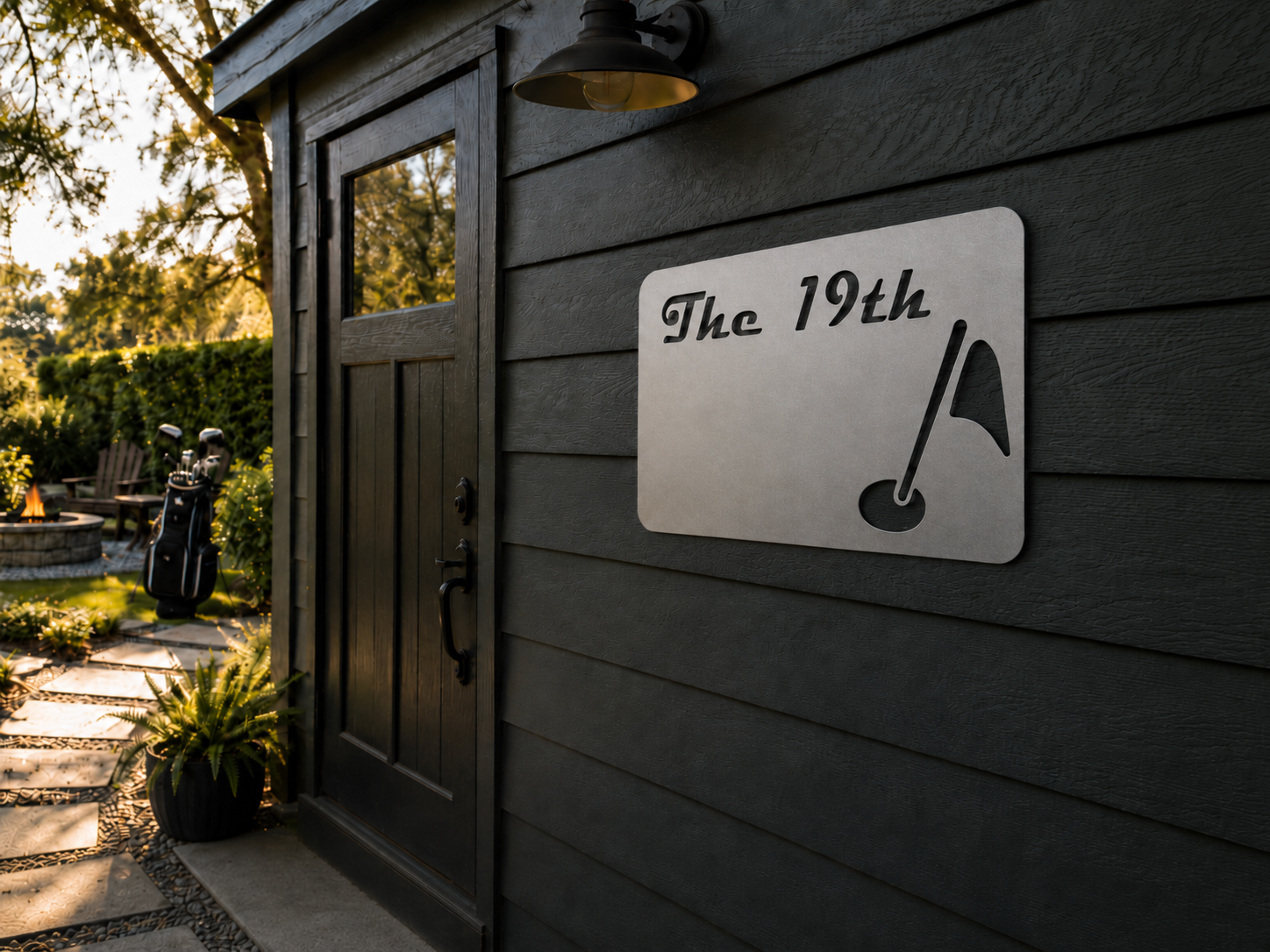 Exterior view of a dark wooden house with a door and a sign that reads 'The 19th' with a golf flag and ball icon. A golf bag with clubs is near a fire pit, with outdoor seating and plants visible in the background.