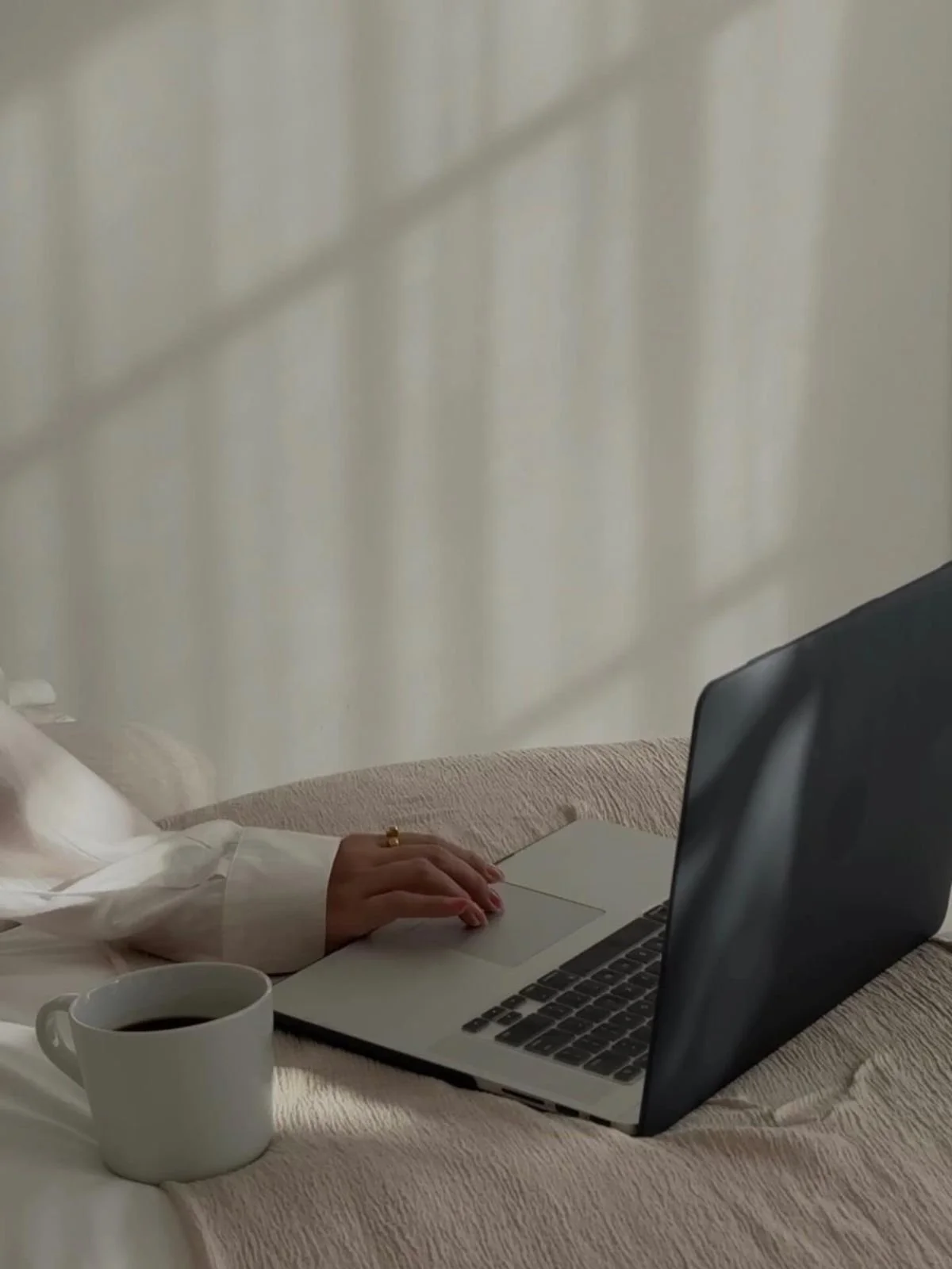 Eine Person arbeitet an einem MacBook Laptop, der auf einem Bett liegt, neben einer weißen Kaffeetasse, mit einem weißen Oberteil und einer goldenen Ring an der Hand.