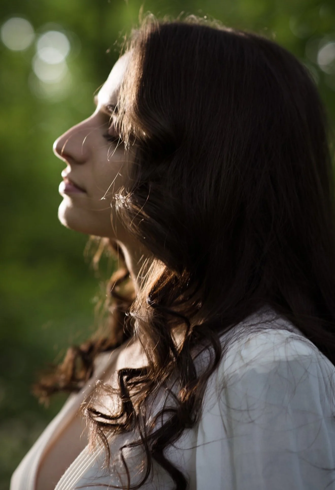 Das Bild zeigt eine Frau in Seitenansicht mit geschlossenen Augen, die in einem grünen, sonnendurchfluteten Außenbereich steht.