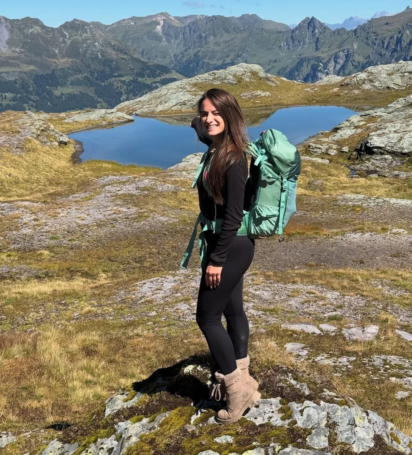 Eine junge Frau wandert in den Bergen mit einem Rucksack auf dem Rücken, vor einem See und hohen Bergen im Hintergrund.