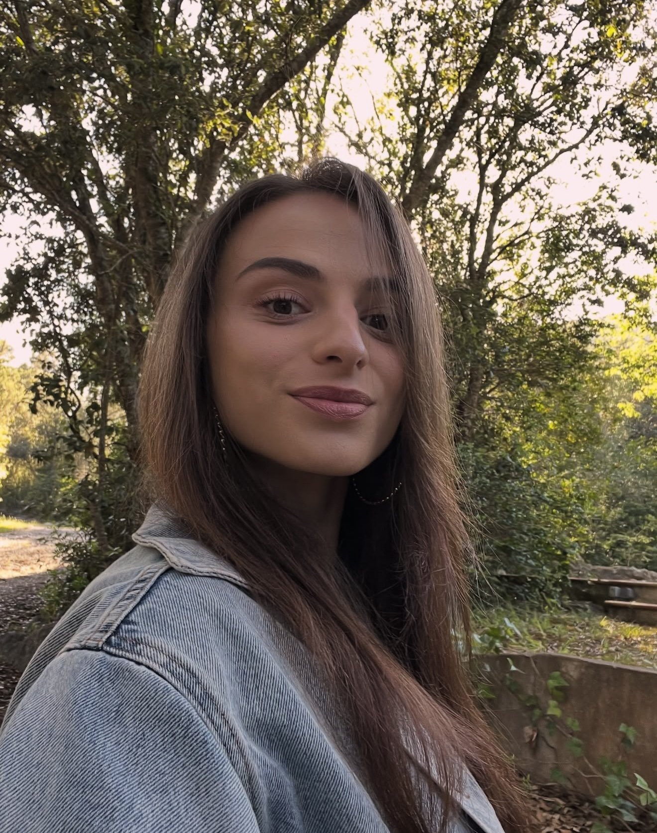 Ein junges Mädchen mit braunen Haaren und Lippenstift, das draußen vor Bäumen steht und in die Kamera lächelt.