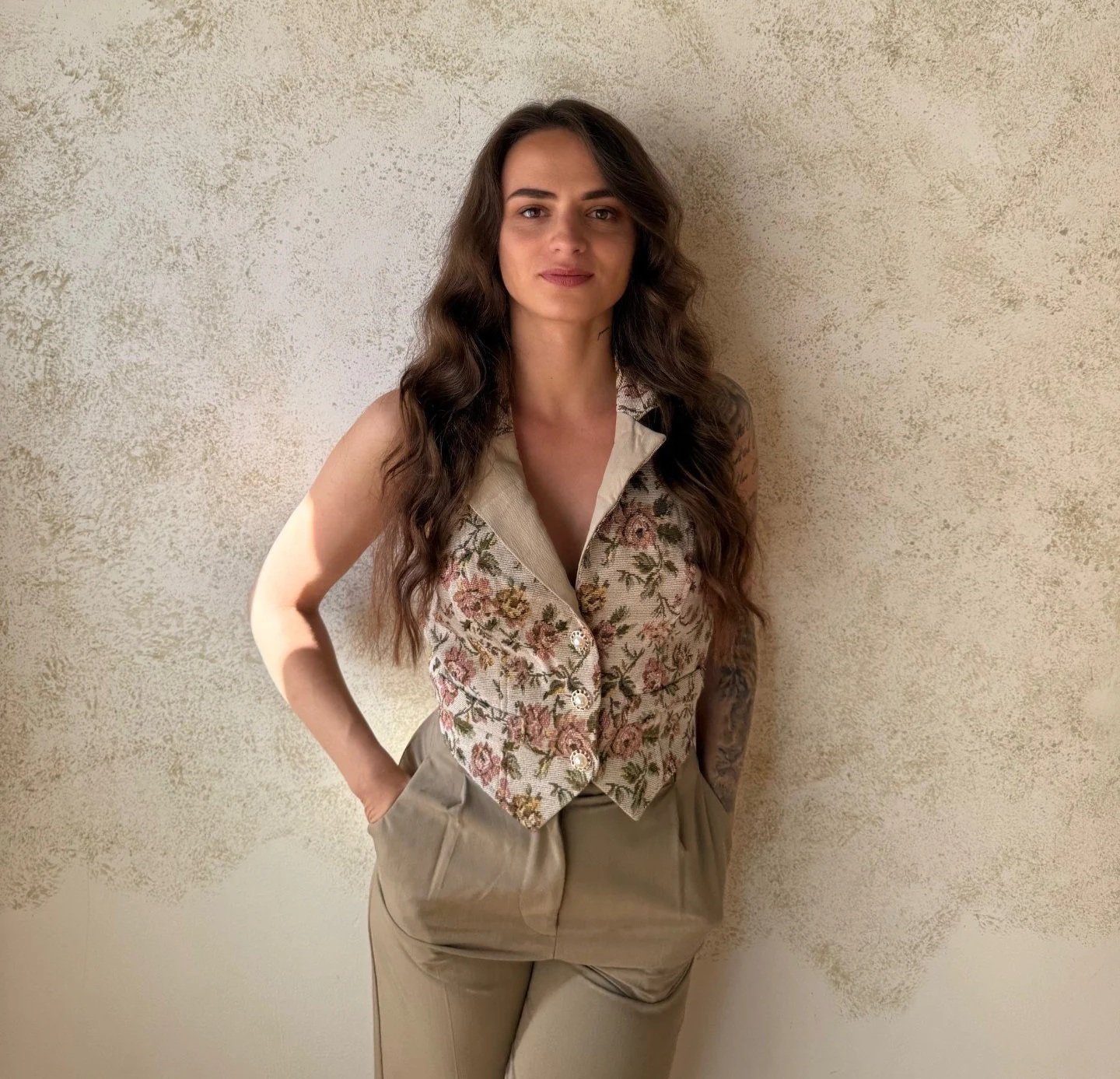Eine Frau mit longem, dunkelbraunem, gewelltem Haar steht vor einer hellbeigen Wand mit strukturierter Oberfläche, trägt eine ärmellose Weste mit Blumenmuster und beige Hosen, schaut in die Kamera und hat die Hände in den Taschen.