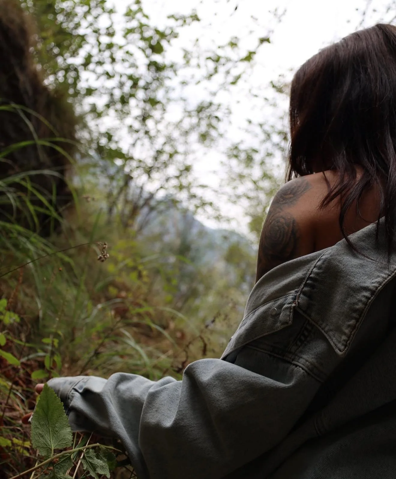 Nahaufnahme einer Person mit Dunklem Haar, tätowiertem Arm und grauer Jacke in einem grünen Waldgebiet.
