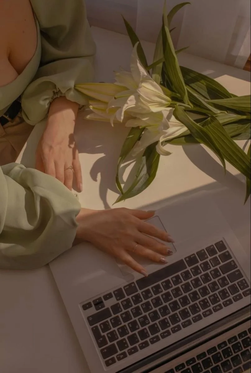 Hand mit Nagellack liegt auf Laptop-Tastatur neben weißen Lilien auf Tisch.