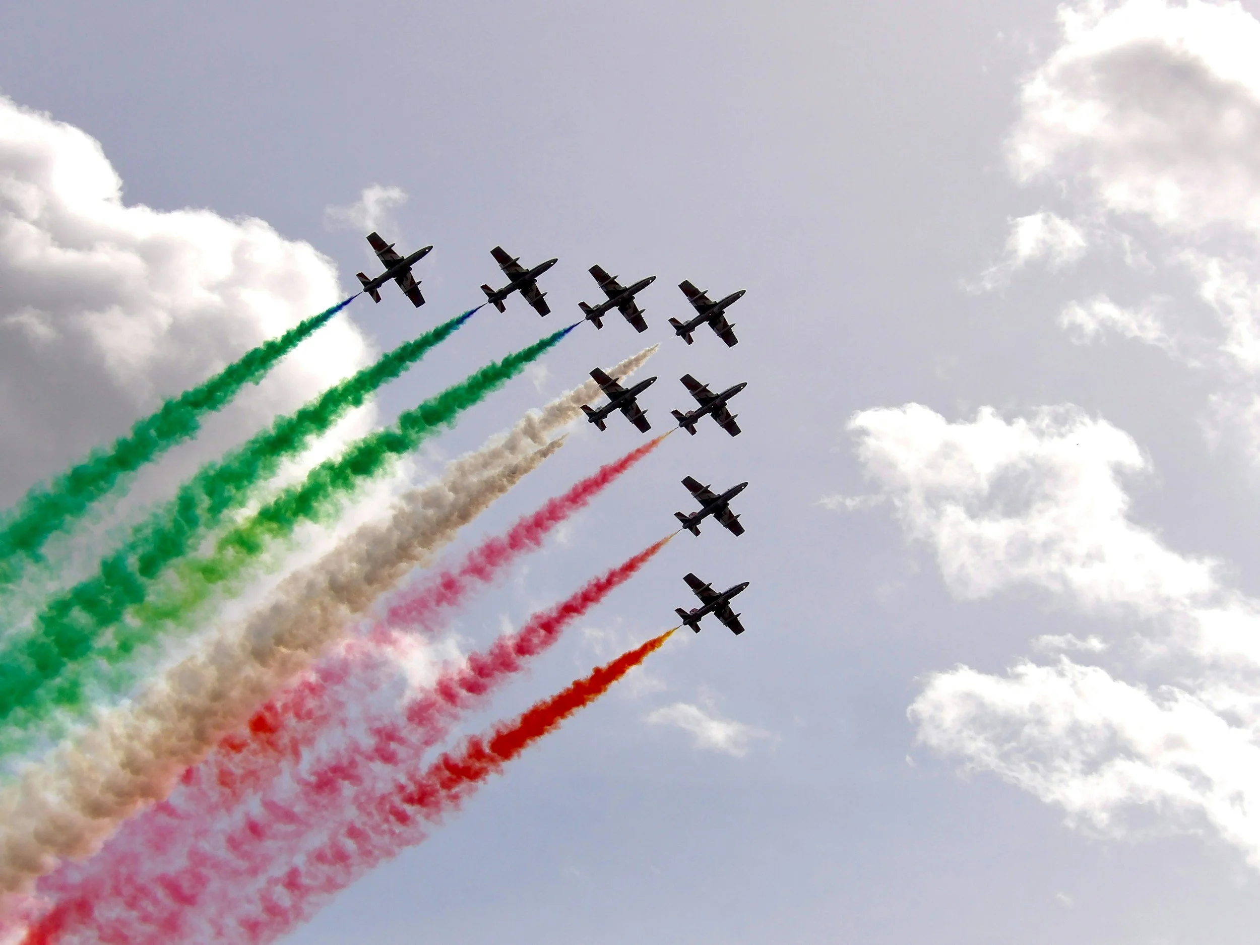 Nine airplanes flying in formation with colored smoke trails in green, white, red, and orange, against a cloudy sky.
