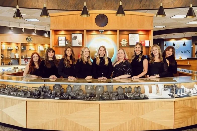 Seven women standing behind a jewelry display counter in a store, smiling at the camera.