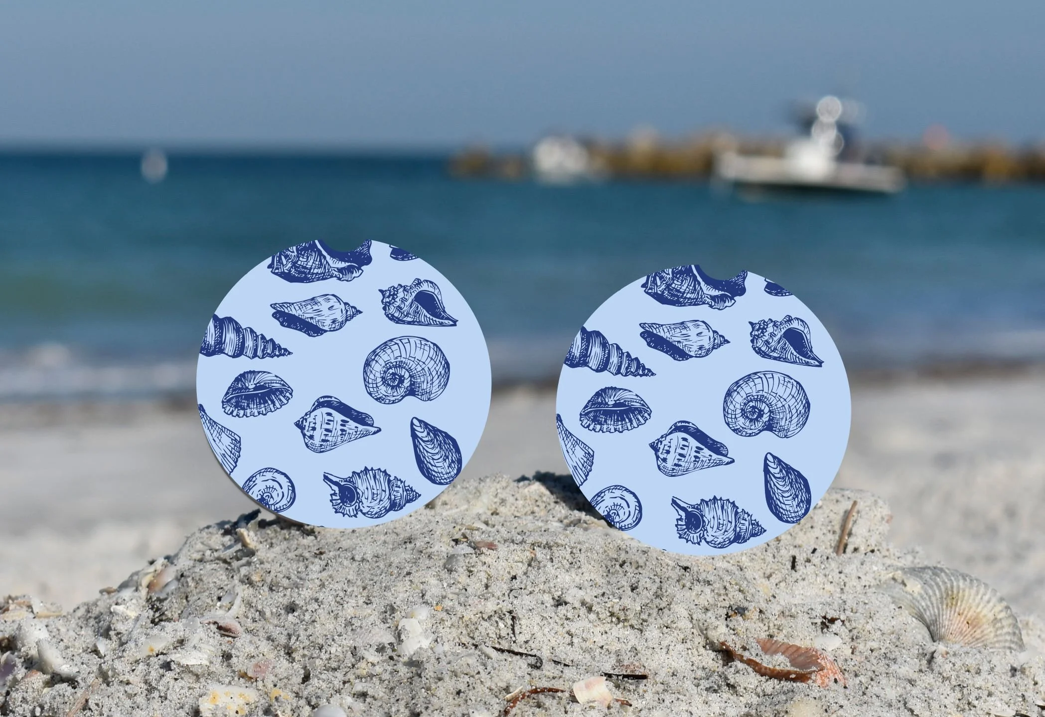 Beach scene with two blue round objects decorated with seashell illustrations, placed on sandy shore with shells, and the ocean with boats in the background.