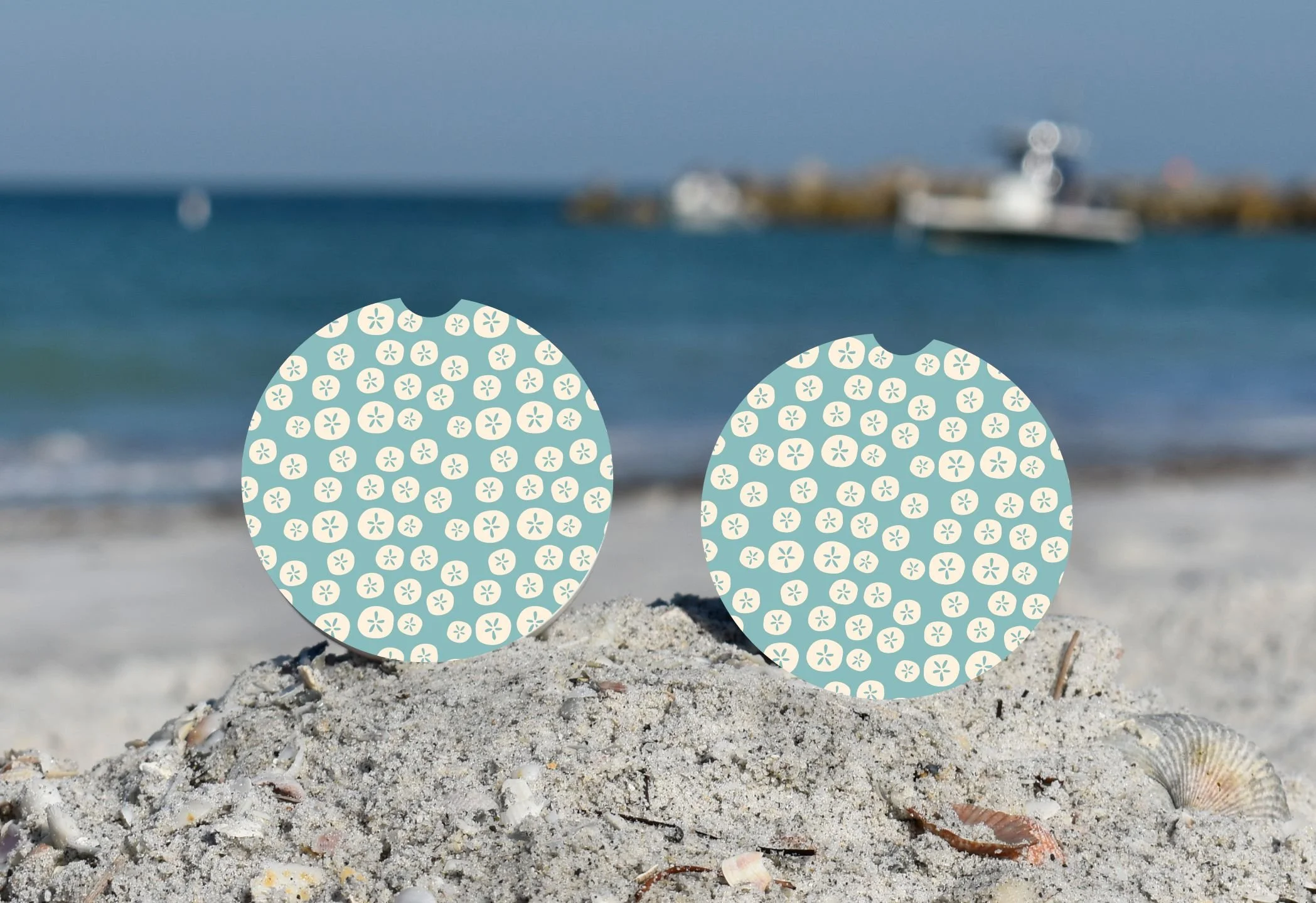 Decorative face masks with blue background and white small flower patterns placed on sandy beach with seashells, ocean, and boats in the blurred background.