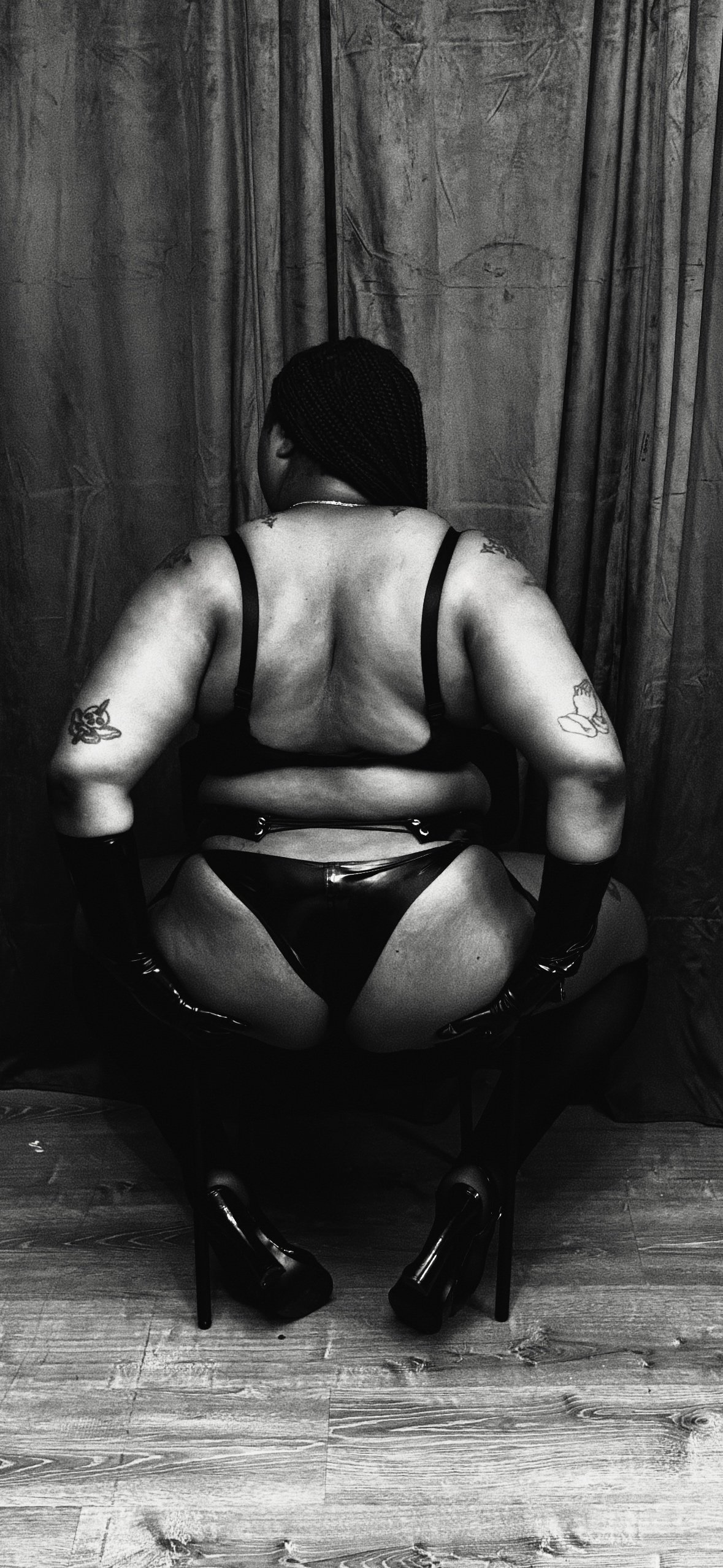 Black woman with braided hair and tattoos on arms, sitting backwards on a chair, wearing black lingerie, latex underwear, and elbow length gloves, with curtains in the background.