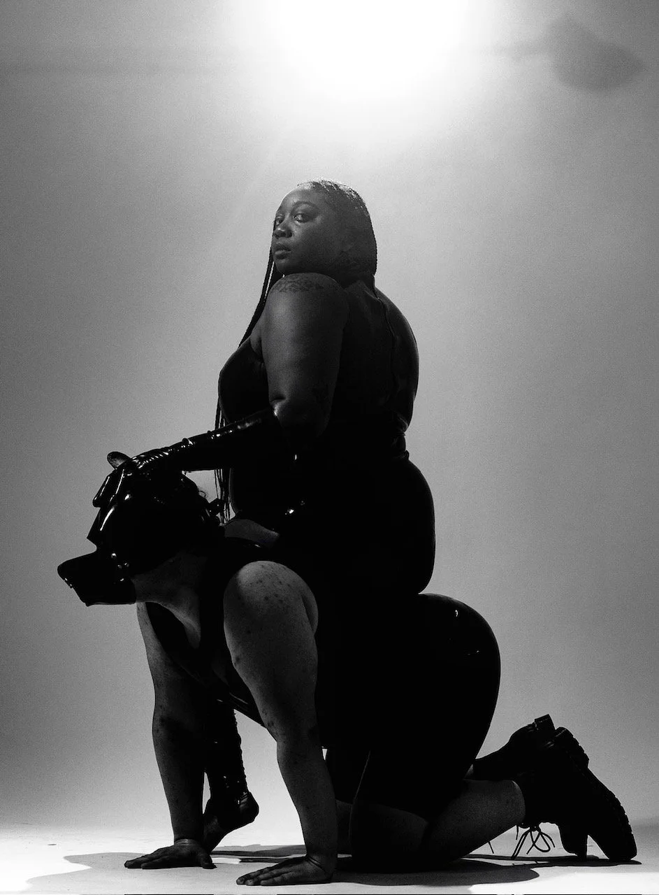 A black and white photo of a man in a latex outfit and dog mask on his hands and knees with a woman in a latex outfit sitting on his back, posing in a studio with a plain background and soft lighting.