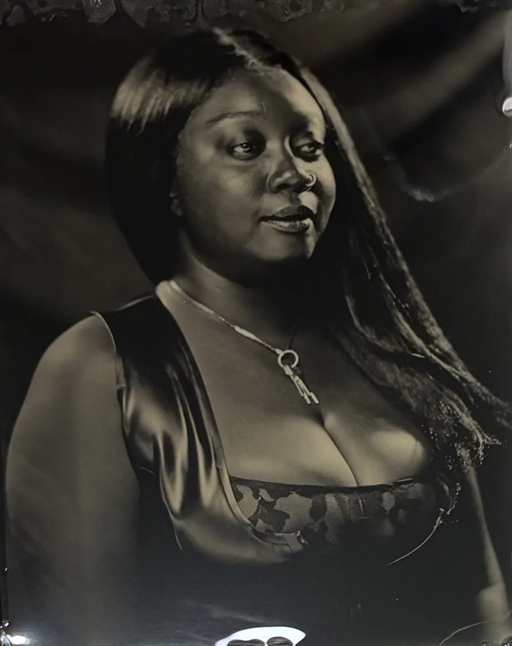 Black and white portrait of a woman with long black hair, wearing a sleeveless top with lace details and a necklace with a key pendant, smiling slightly.