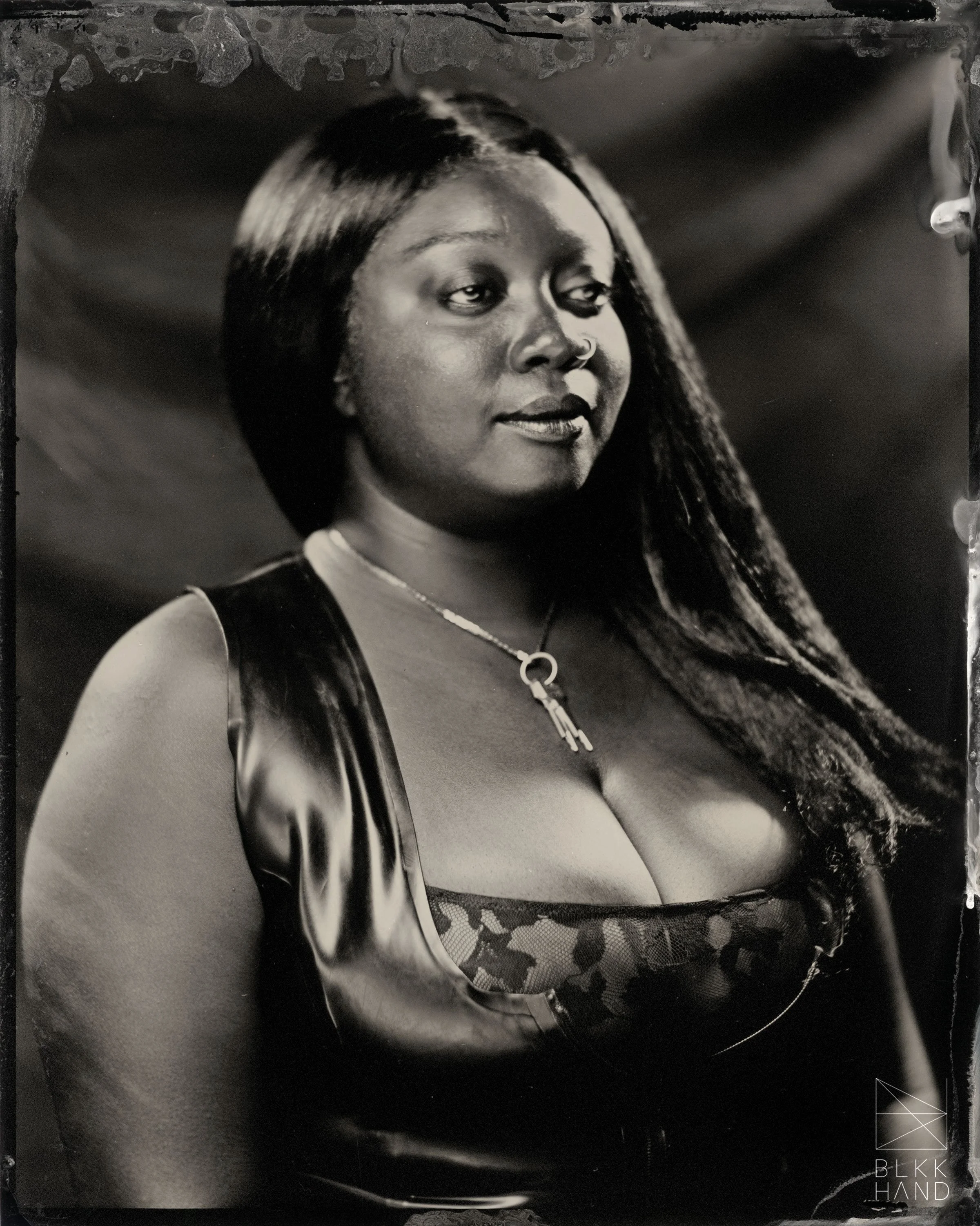 Ebony African American Dominatrix wearing a latex and lace top with a visible chastity key around her neck. Her hair is long, black, and straight, and her face is depicted as smirking and looking to the right.