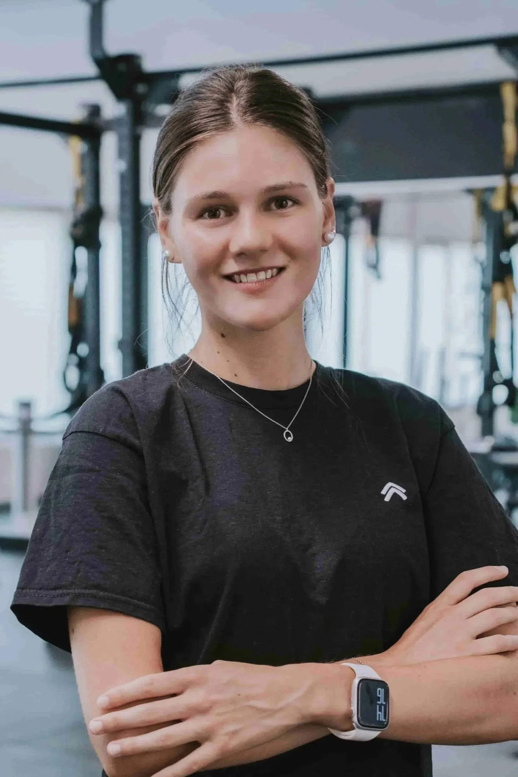 Junge Frau im Fitnessstudio trägt ein schwarzes T-Shirt, trägt eine Uhr am Handgelenk und lächelt in die Kamera.