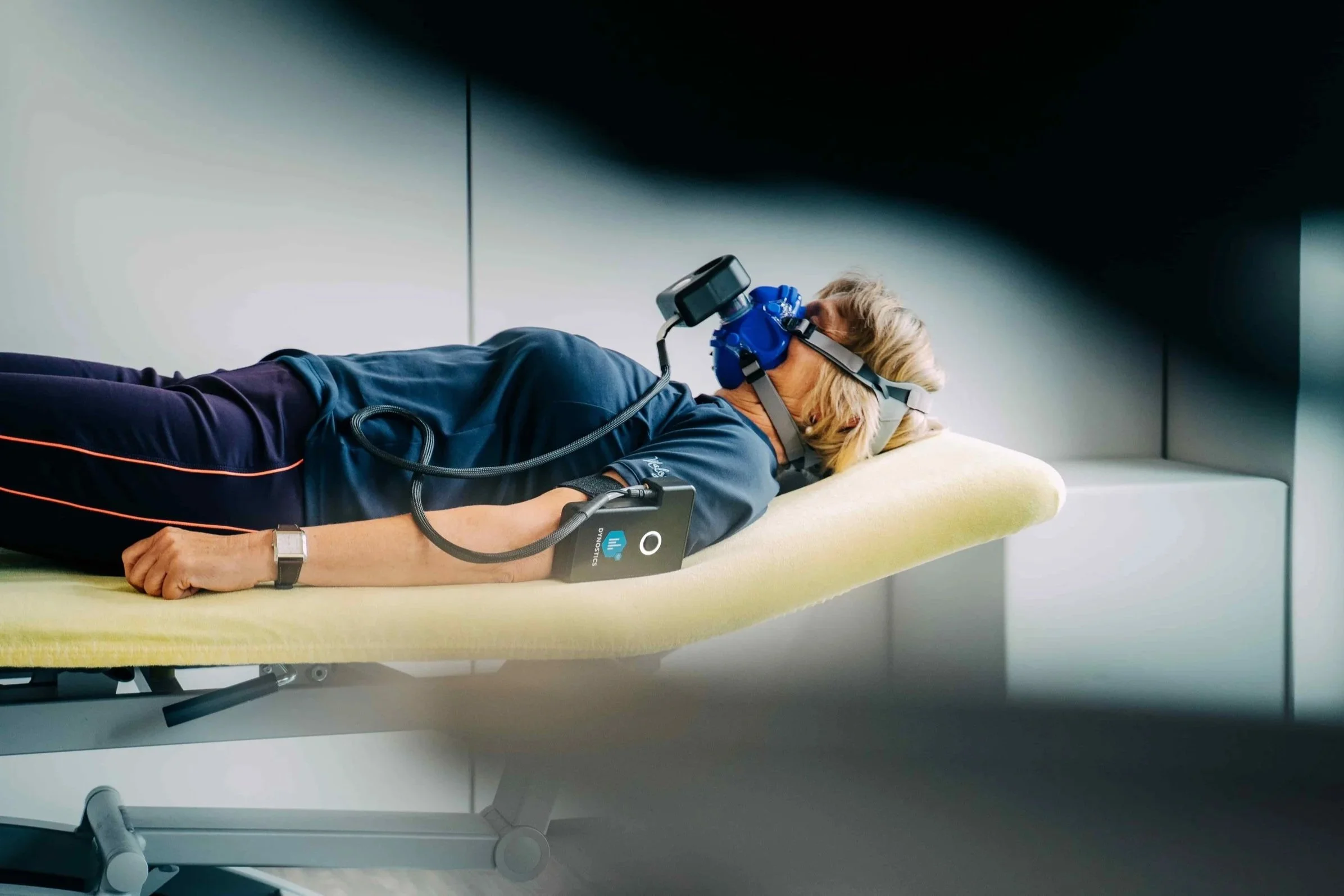 Eine Frau liegt auf einer Untersuchungsliege mit einer Neurorehabilitationsmaschine, trägt eine Kopfhaube mit Sensoren, und ist an ein medizinisches Gerät angeschlossen, in einem klinischen Raum.