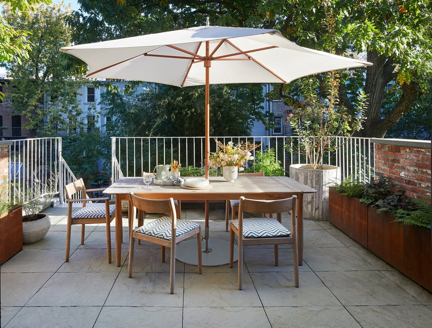 Empty outdoor patio with round table, four chairs with patterned cushions, white umbrella, potted plants, and surrounding greenery