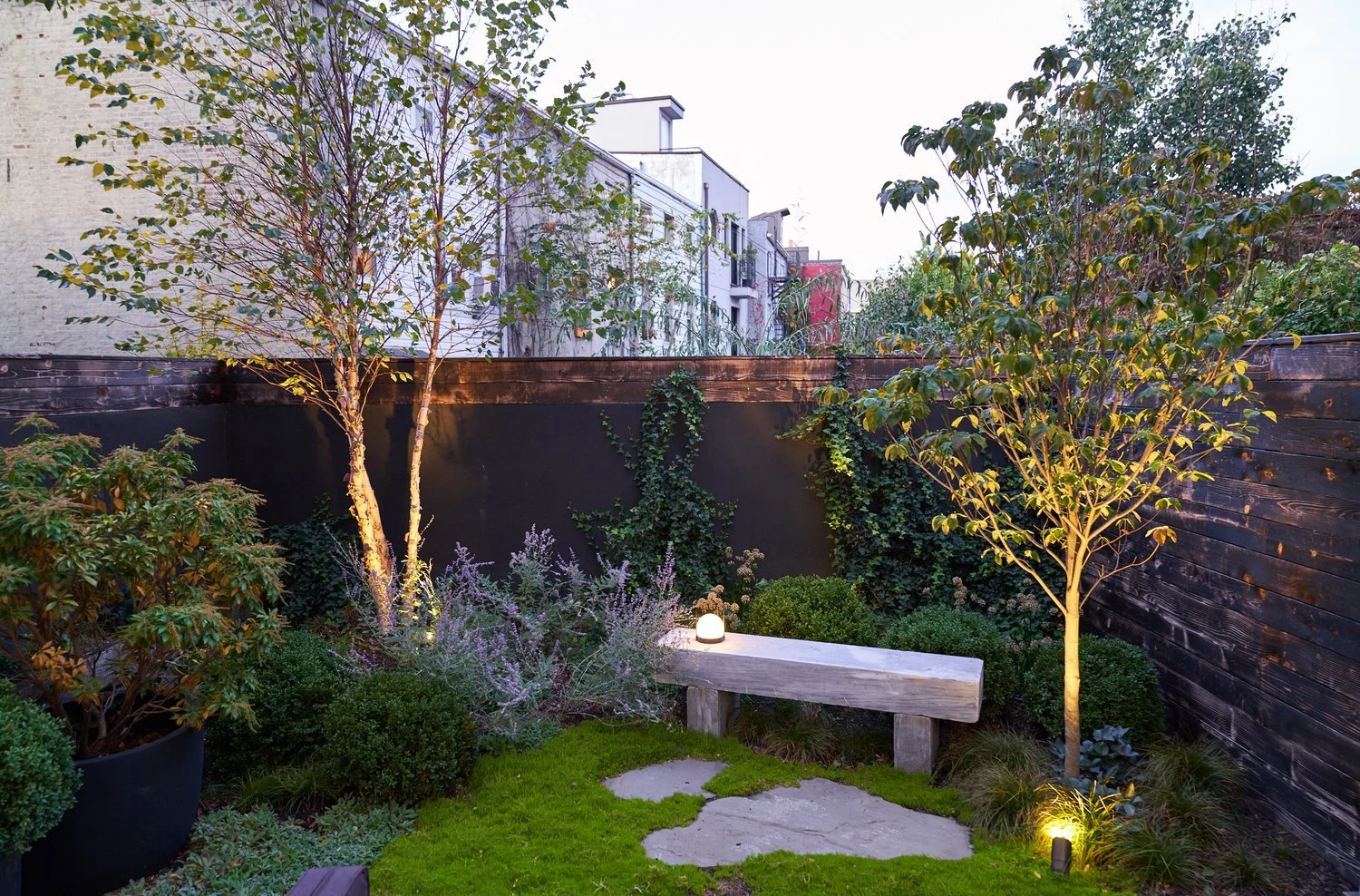A cozy backyard garden at dusk with two lit trees, a stone path, various plants, and a wooden bench.
