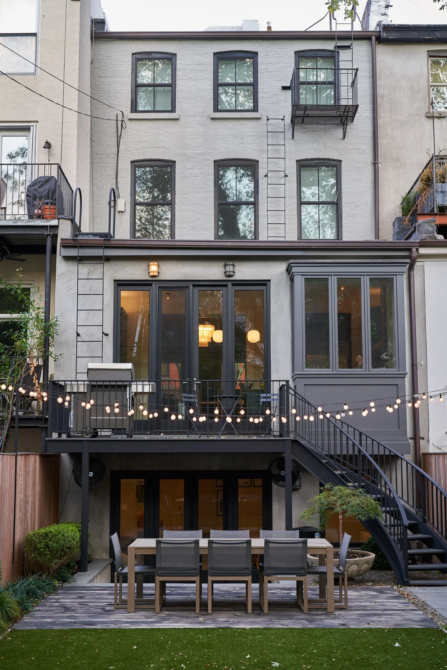 Backyard patio with outdoor dining table and chairs, string lights, and a multi-story building with windows and balconies.