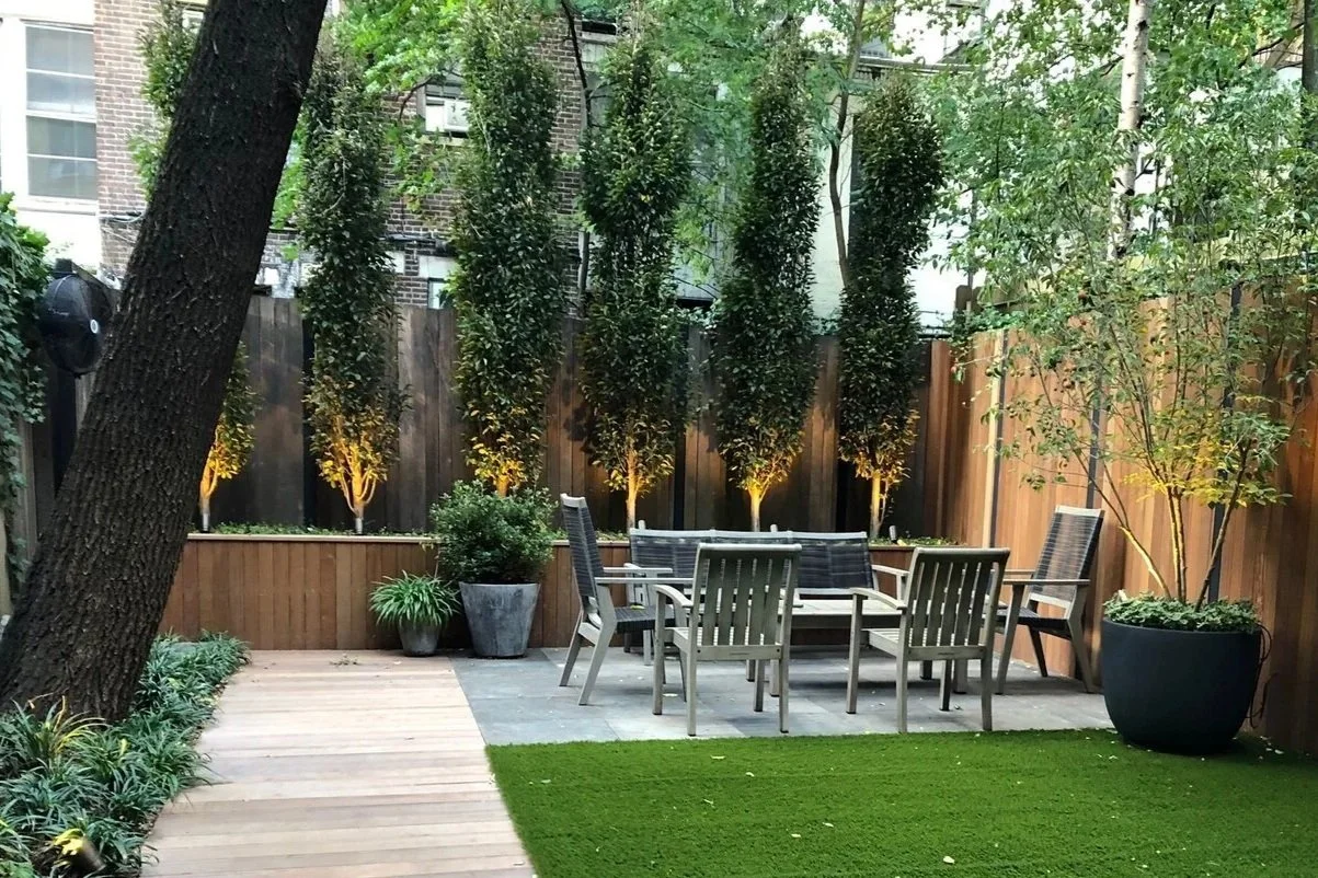 A backyard patio area with outdoor furniture, including chairs and a table, surrounded by potted plants and trees with a wooden fence in the background, illuminated by ground lights.