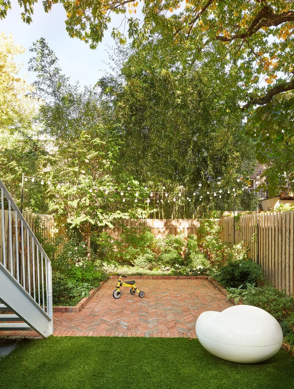 Backyard with a brick patio, green trees, string lights, a small yellow tricycle, a modern white outdoor lounge chair, and a grassy area.