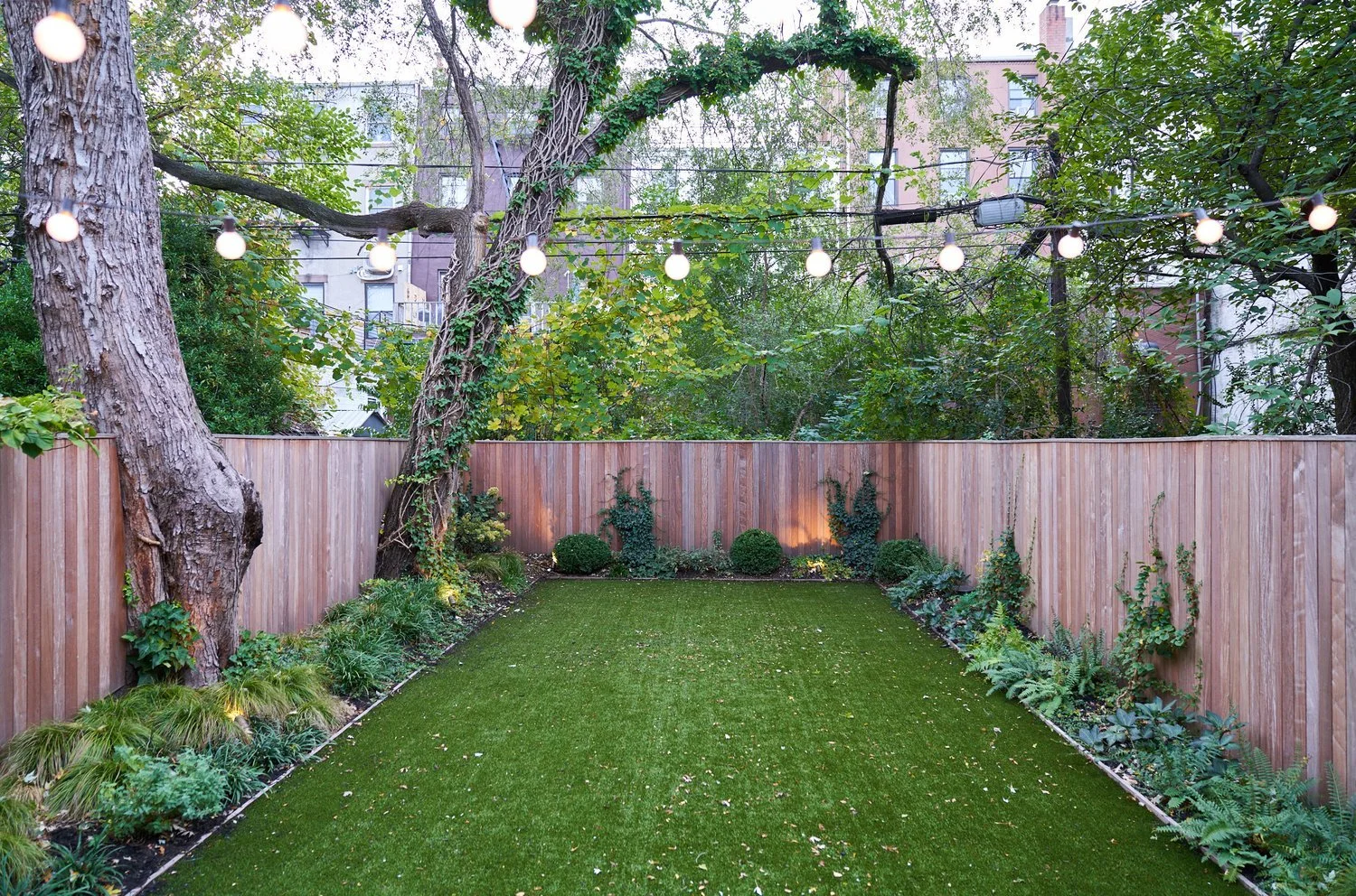 A backyard garden with a wooden fence, large leafy trees, string lights overhead, and a lush green lawn bordered by plants and shrubs.