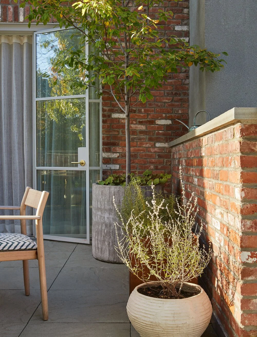 Outdoor patio with potted plants, a glass door with curtains, a wooden chair, and brick wall.