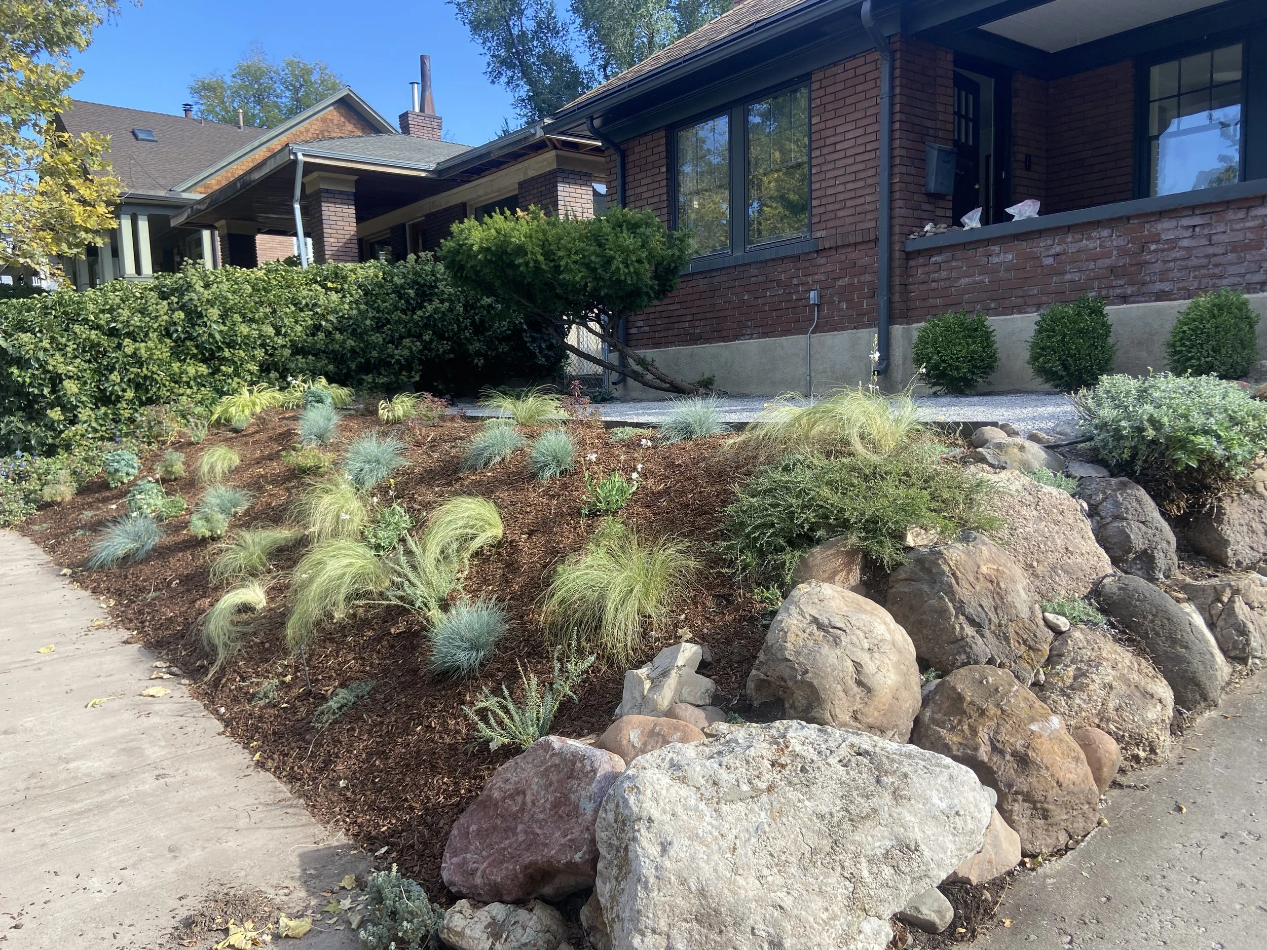 Residential front yard with well-maintained garden, rocks, shrubs, and a brick house with large windows and a porch.