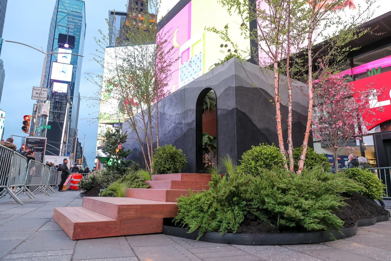 Urban scene with a small landscaped area featuring trees, bushes, and pink blossoms, surrounded by metal barriers, with tall skyscrapers and digital billboards in the background.
