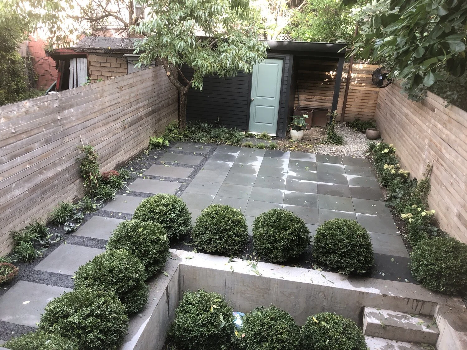A small backyard with stone tiles, a wooden fence, and landscaped bushes along the edges. There is a small black shed with a light blue door, a tree, some planters, and a covered seating area in the back corner.
