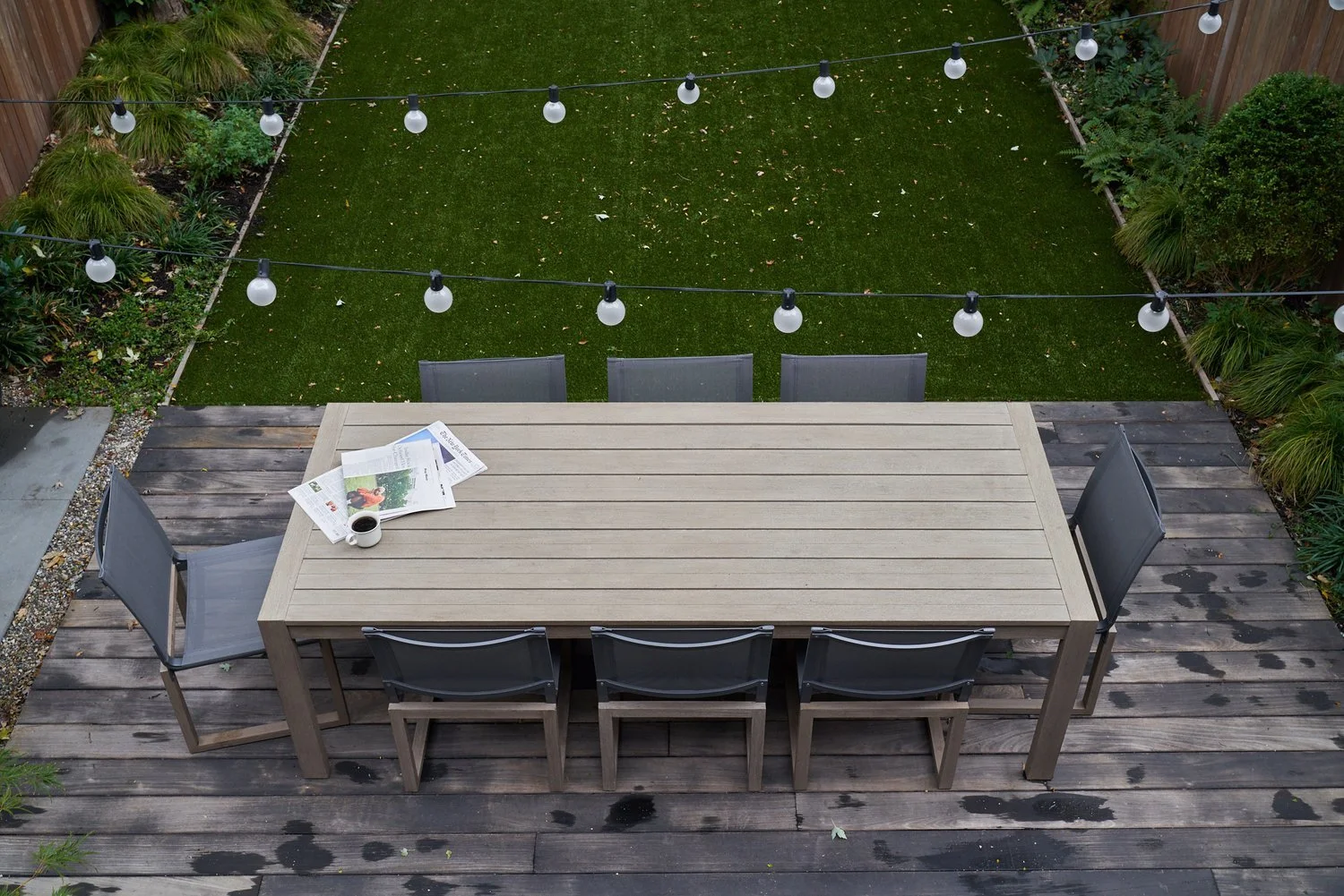 An outdoor dining table with six chairs on a wooden deck, with magazines and a cup of coffee, overlooking a grassy yard with string lights overhead and garden plants along the edges.