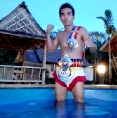 Person standing in a pool wearing a Muay Thai outfit with championship belts, in an outdoor setting with thatched roofs in the background.