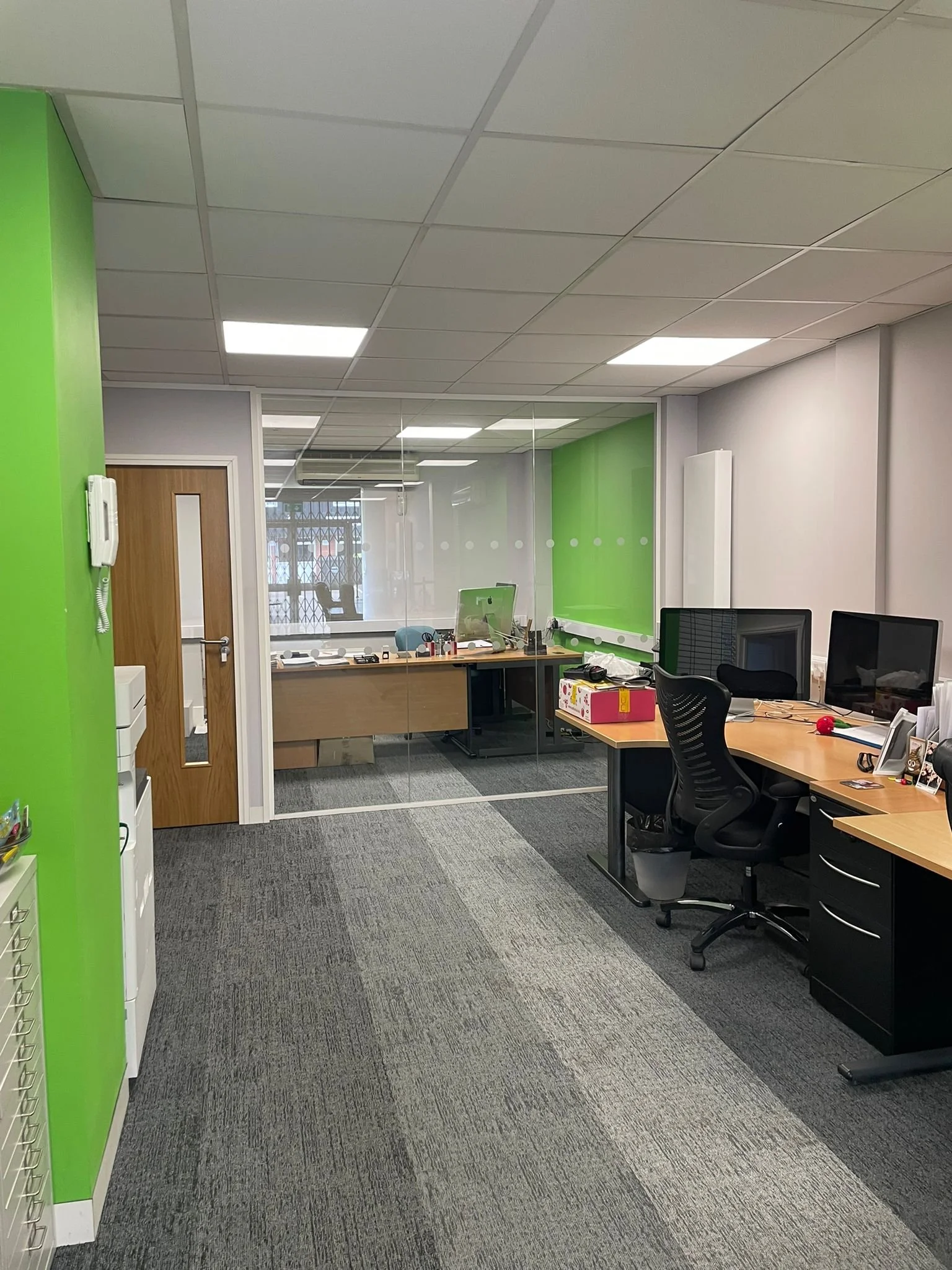 Office space with desks, computers, and a green accent wall, viewed through a glass partition, with a hallway and closed door on the left.