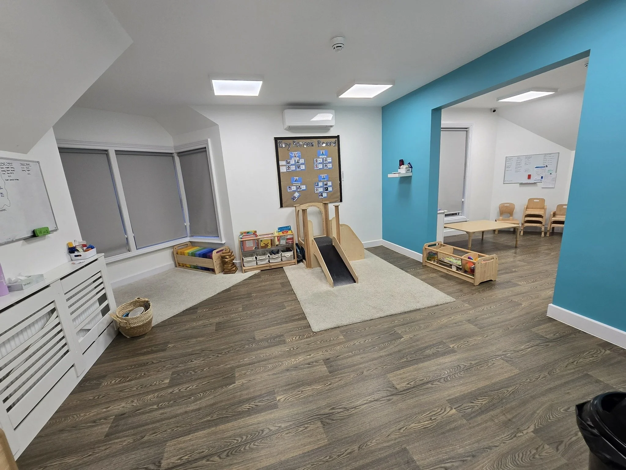 A classroom with white walls and wood flooring, featuring a playhouse slide, a bulletin board, chairs, and a small table.