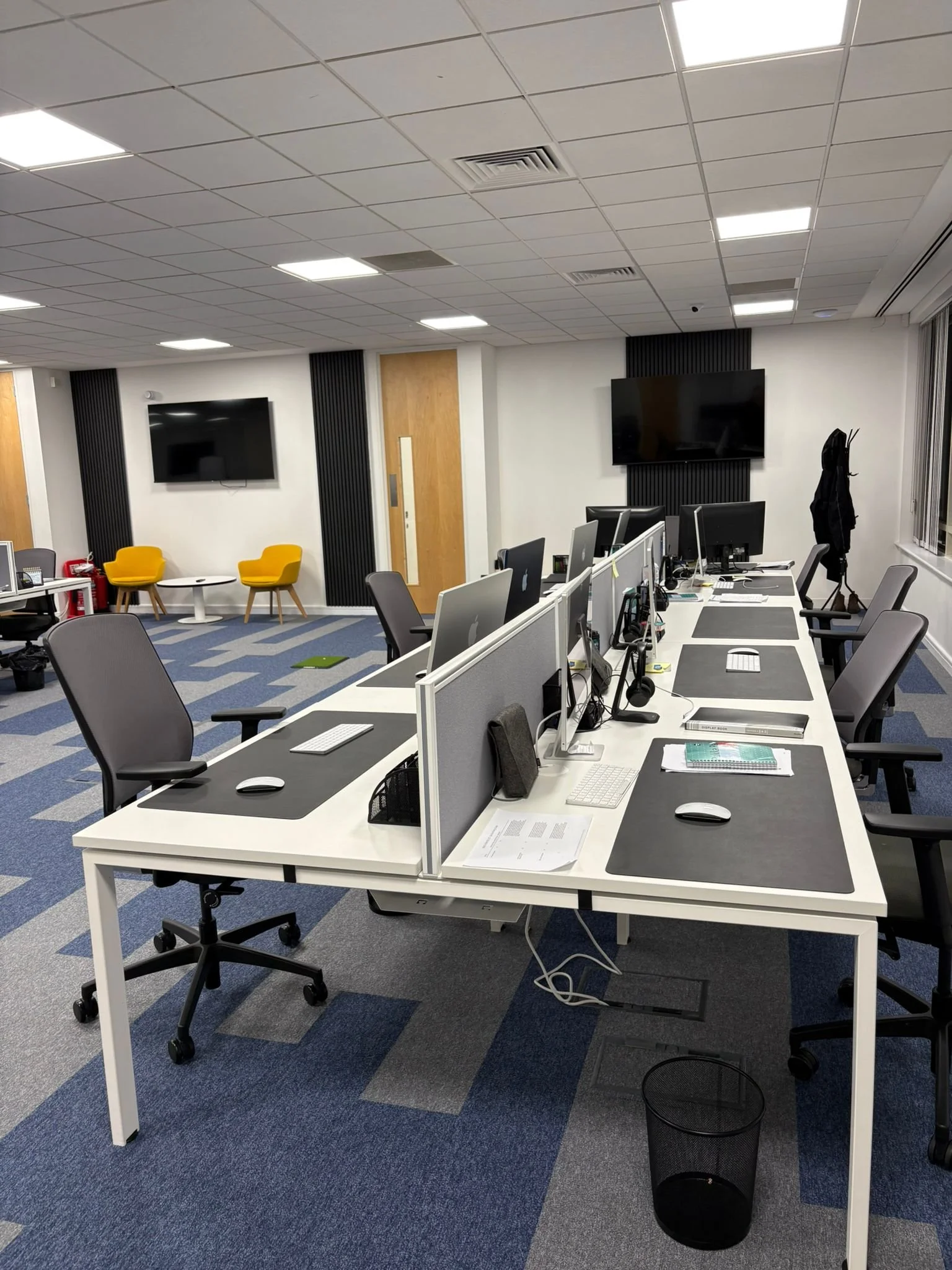 An office space with white desks arranged in a row, each equipped with black desk pads, computers, and office supplies. The room has gray chairs, a blue and gray patterned carpet, and a ceiling with square tiles and fluorescent lighting. In the background, there are two yellow chairs, a small round white table, two large flat-screen TVs on the wall, a wooden door, and a coat rack.