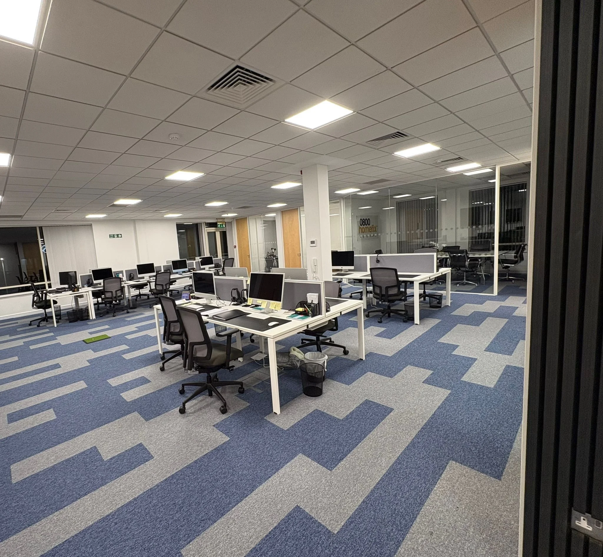 An office space with rows of desks and multiple computer monitors. The room has a patterned blue and gray carpet, white walls, and a drop ceiling with fluorescent lighting. A glass wall separates this office from another area.