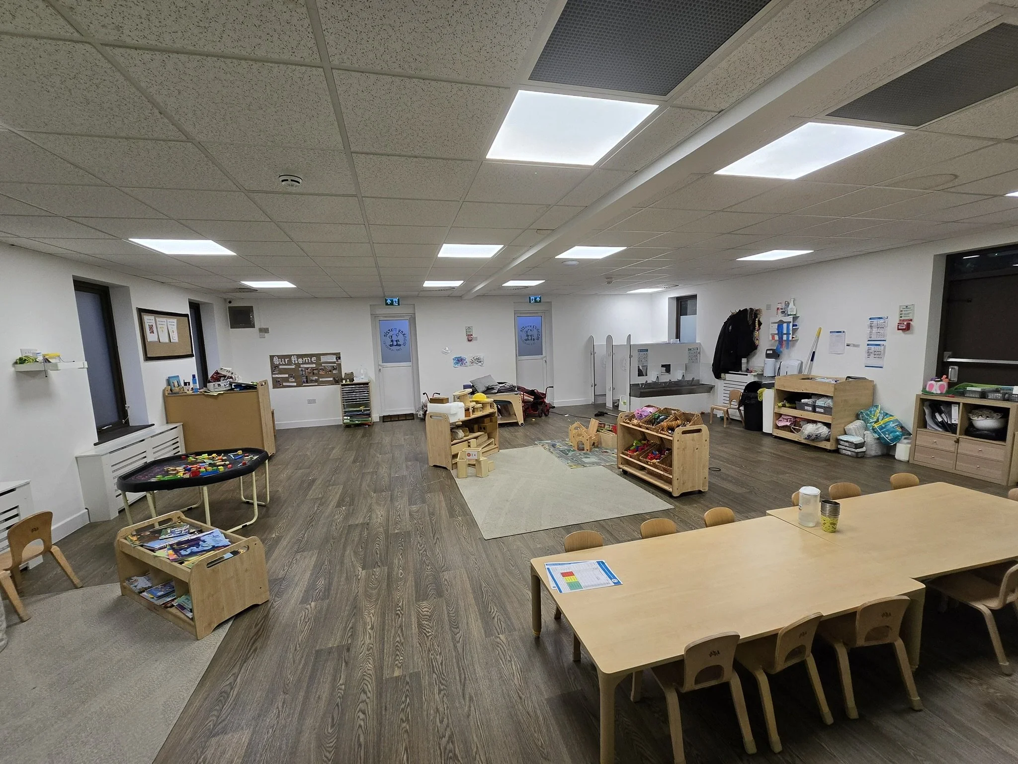 A classroom with a wooden table and chairs in the foreground, scattered toys and books, with a bulletin board, doors, and ceiling lights in the background.