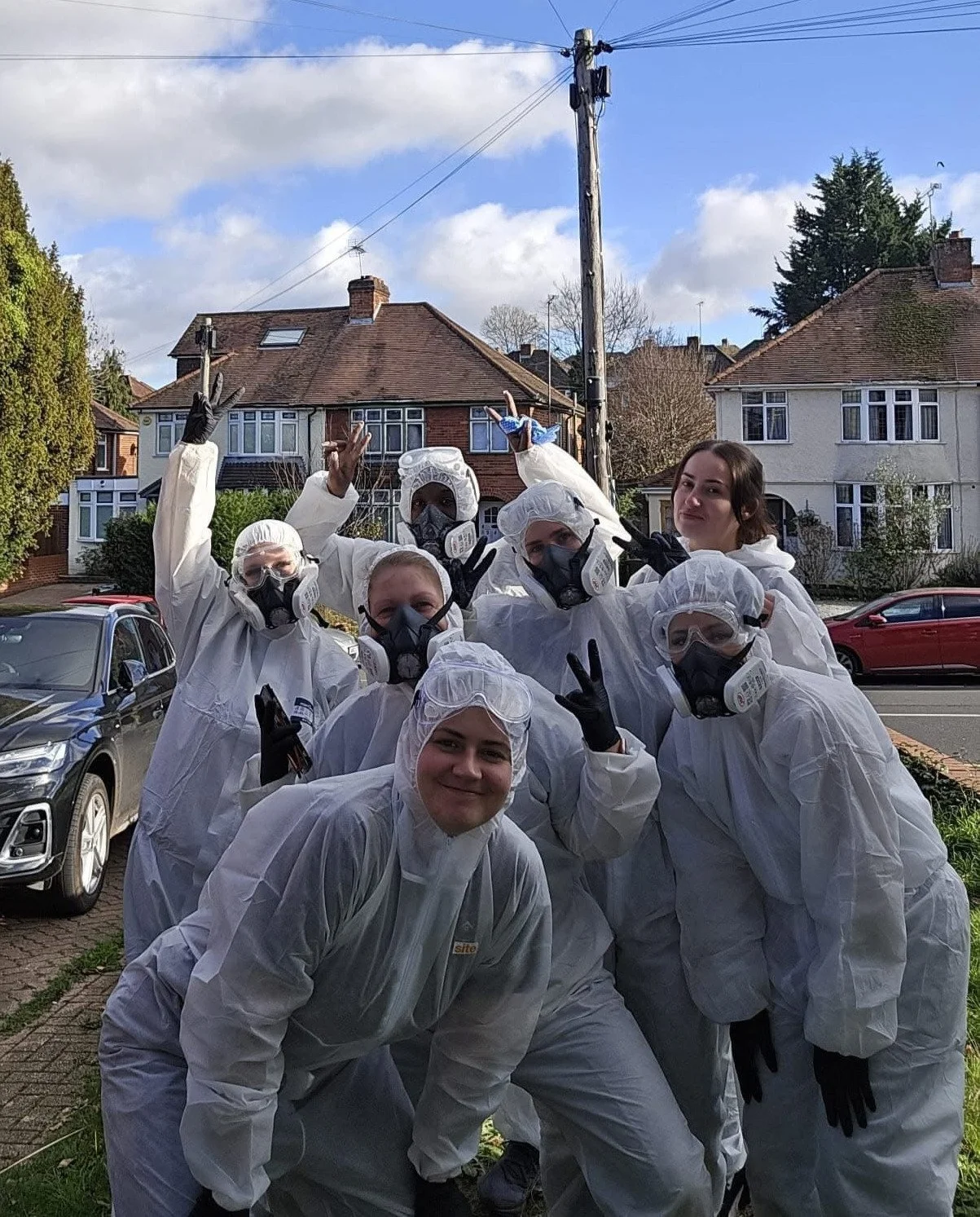 Group of people in white protective suits and masks posing outdoors on a residential street, some making peace signs.