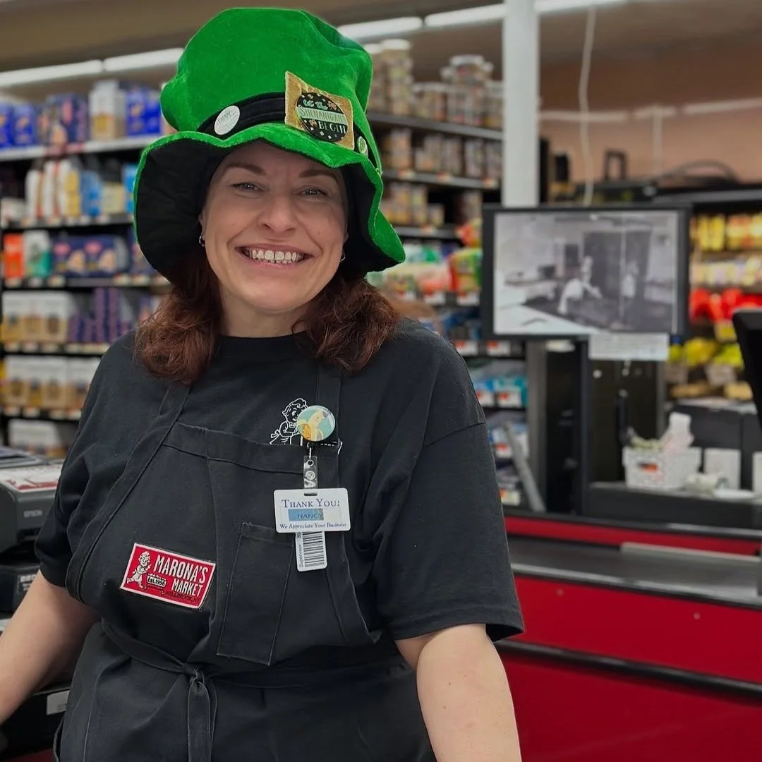 A smiling woman wearing a large green hat and a black uniform stands in a grocery store checkout area. The background includes store shelves stocked with various products and a checkout counter with a POS machine.