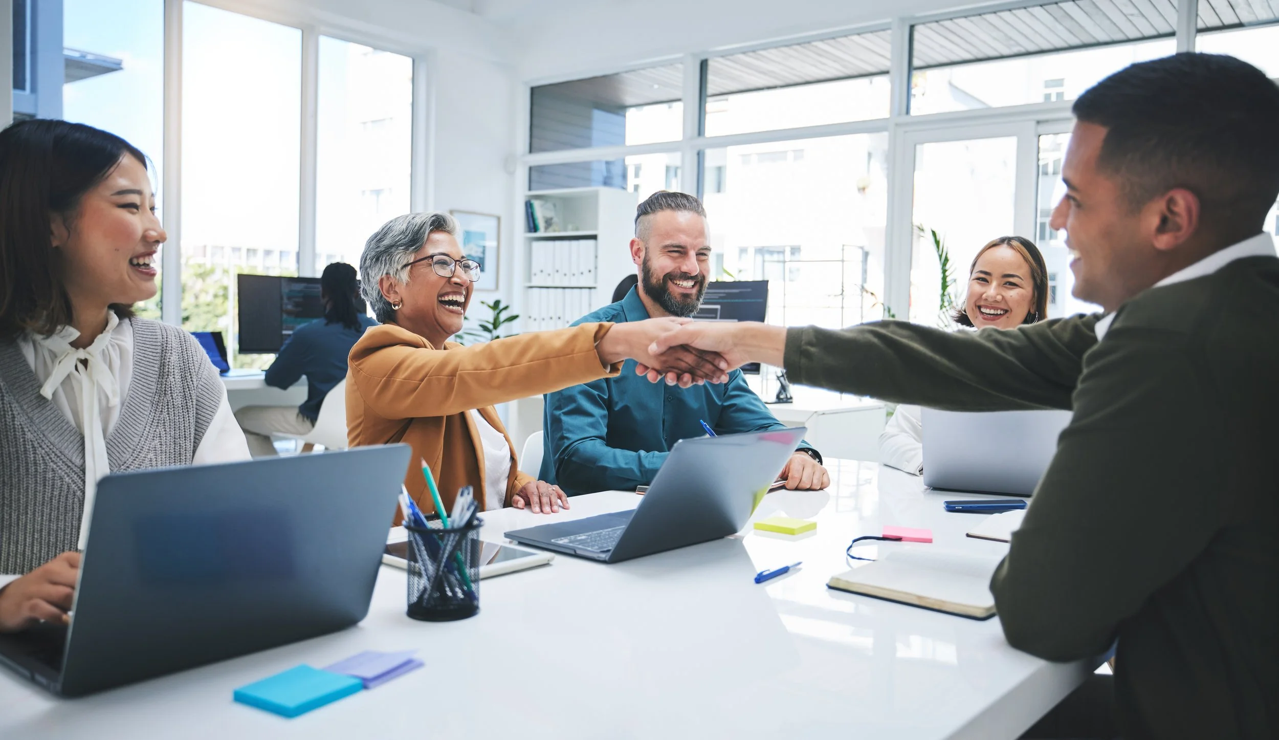 Group of five diverse colleagues in an office, smiling and shaking hands across a table, with laptops and office supplies nearby.