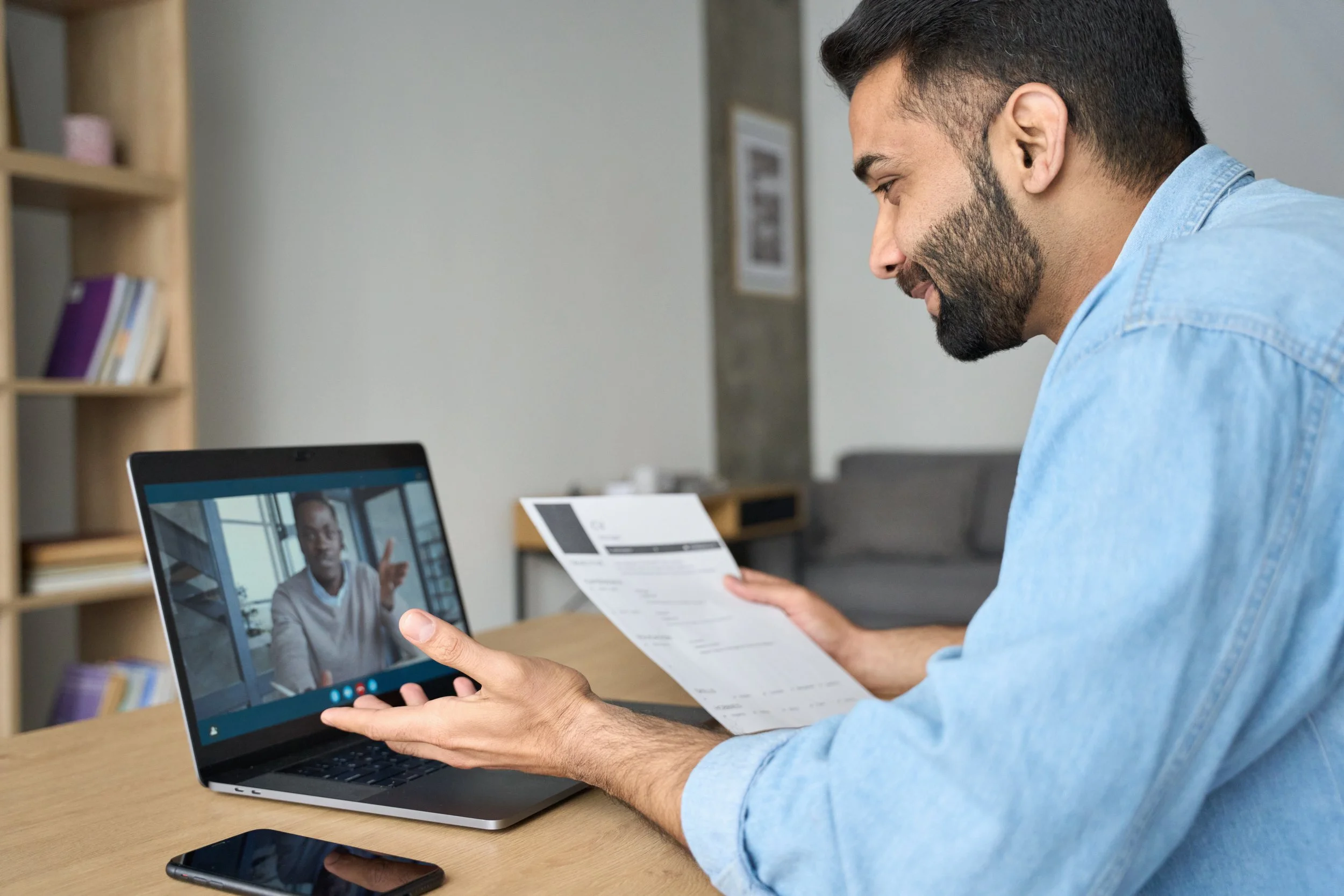A man holding a paper and smartphone, engaged in a video call with a woman on his laptop screen, in a home or office setting.
