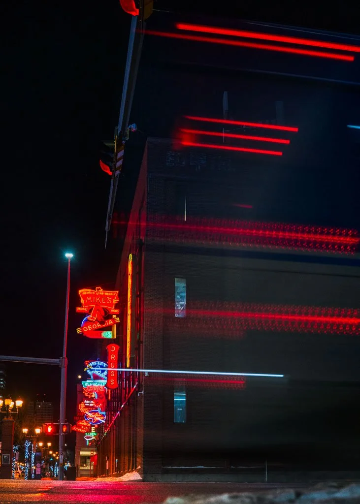 Nighttime city street with neon signs, including one for 'Mike's' and a drug store, with light trails from passing vehicles.