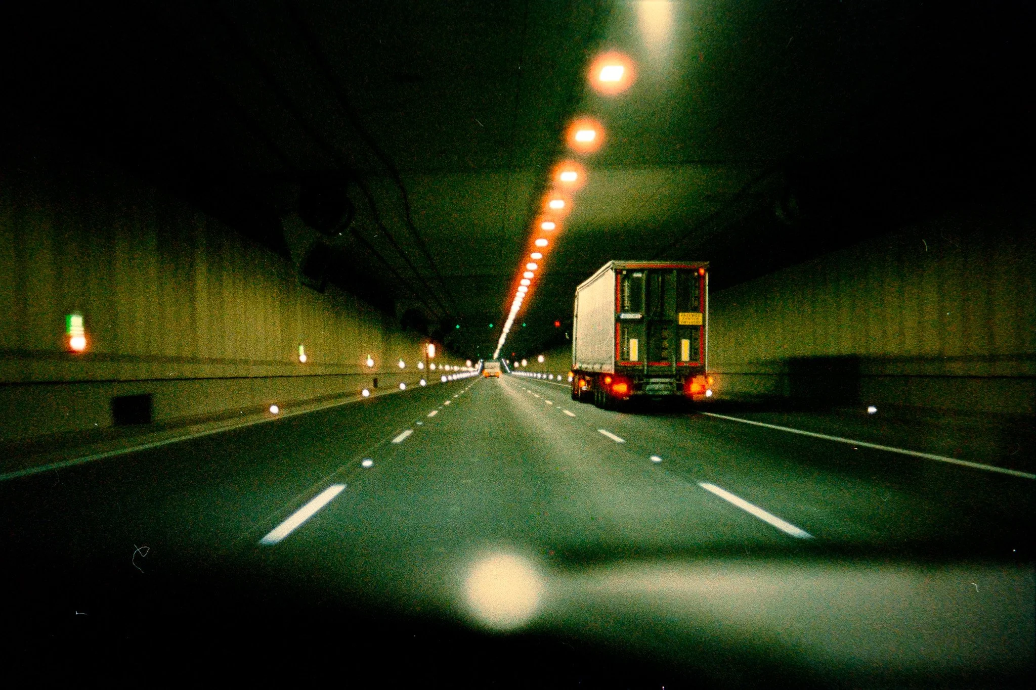 View of a highway tunnel at night with lane markings, vehicle, and lights along the tunnel ceiling.