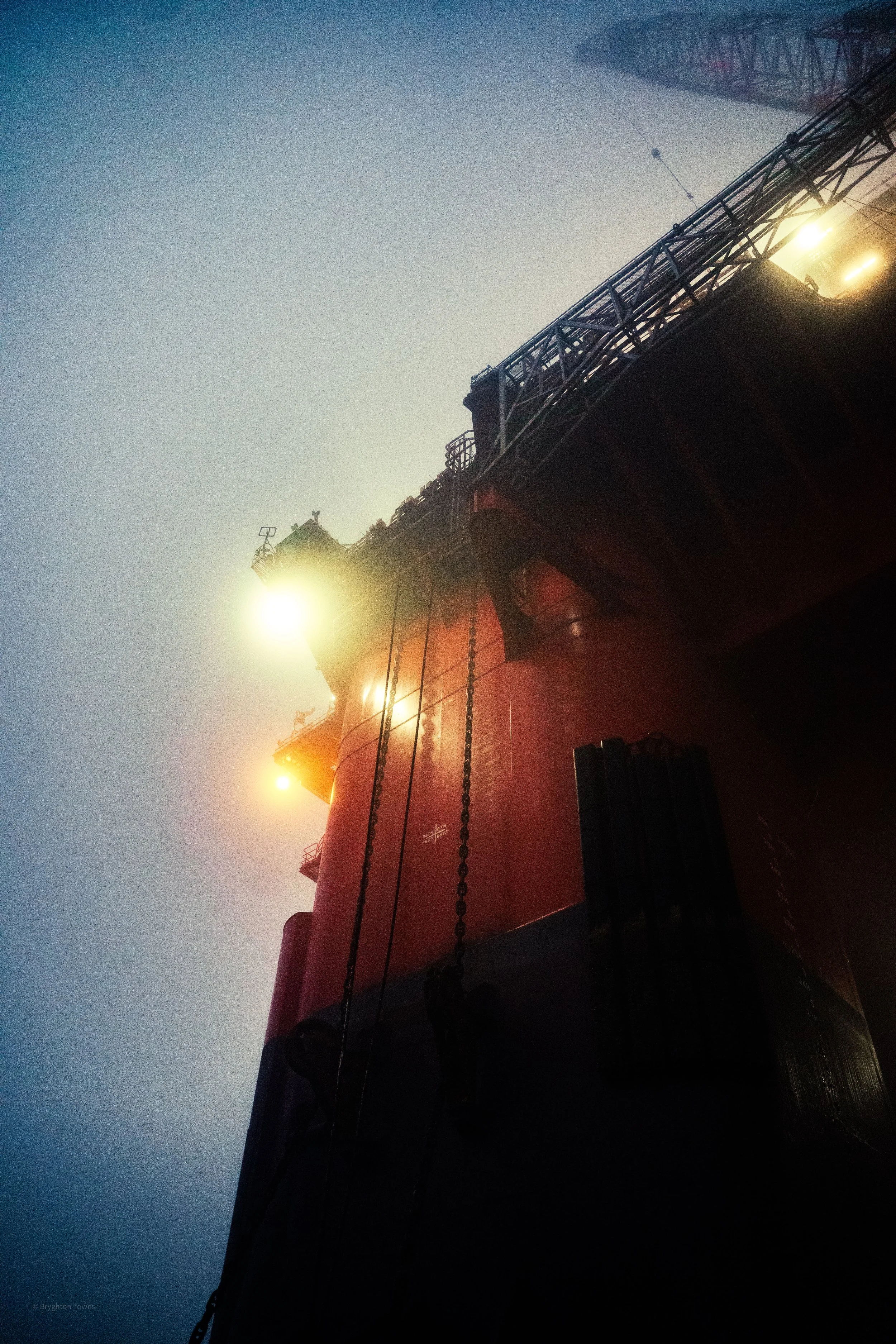Close-up view of an offshore oil rig or platform with bright lights against a misty, blue sky, showing structural details and chains hanging from the platform.