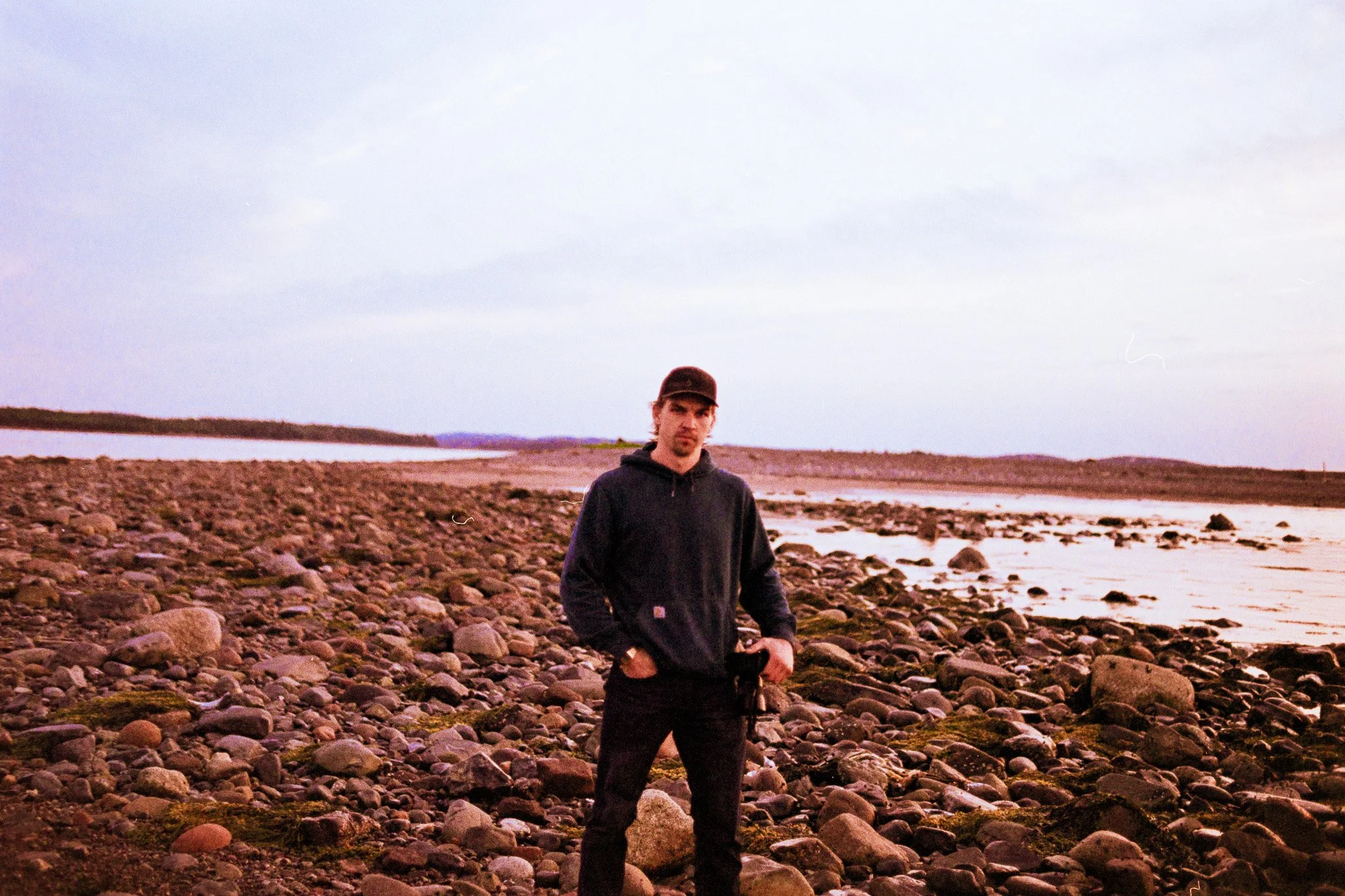 A young man standing on a rocky beach near a body of water, with a cloudy sky overhead.